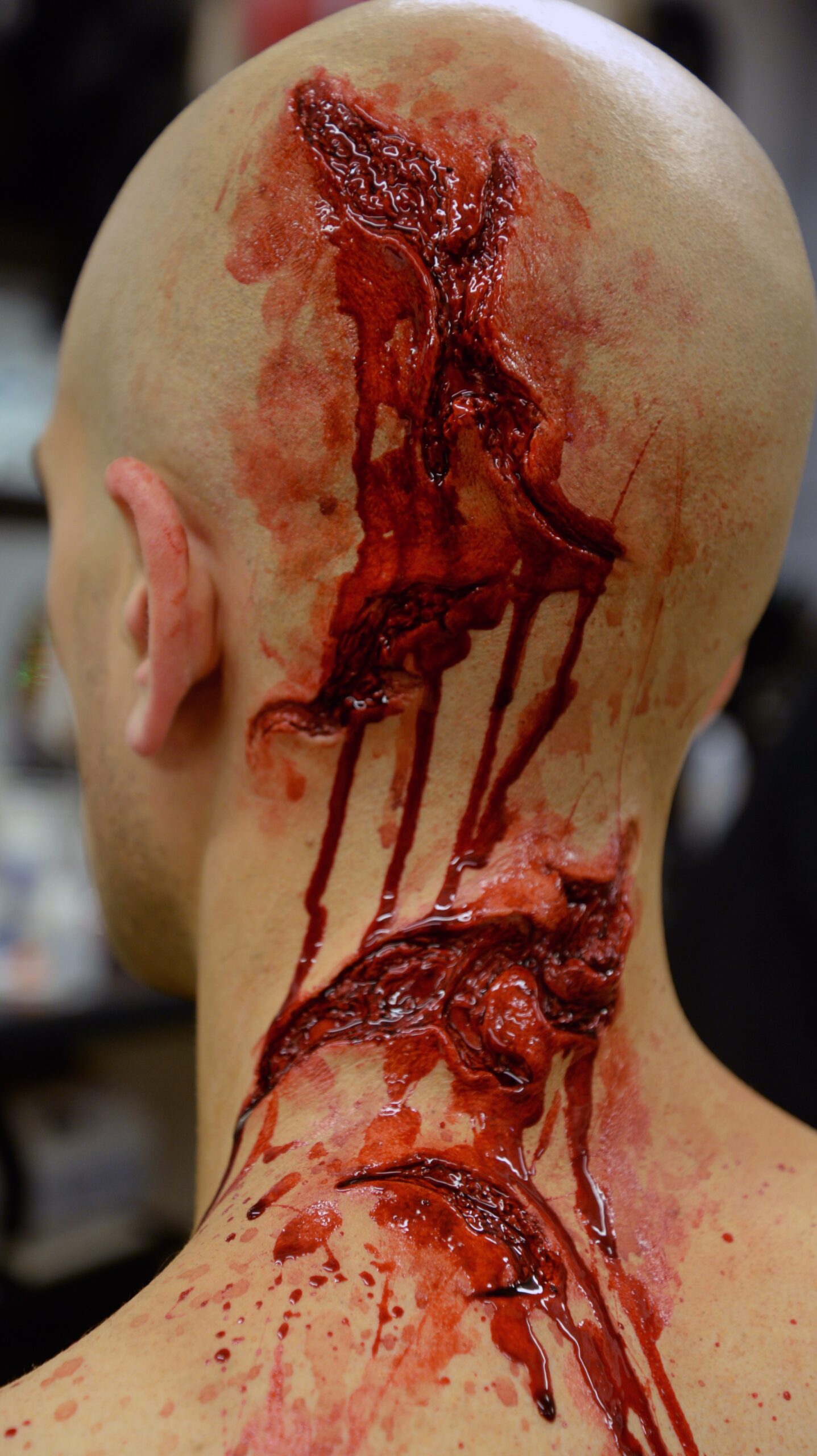A person with a bald head displays realistic-looking, trauma-inspired special effects makeup simulating deep, bloody wounds running from the top of the scalp down the back of the neck.