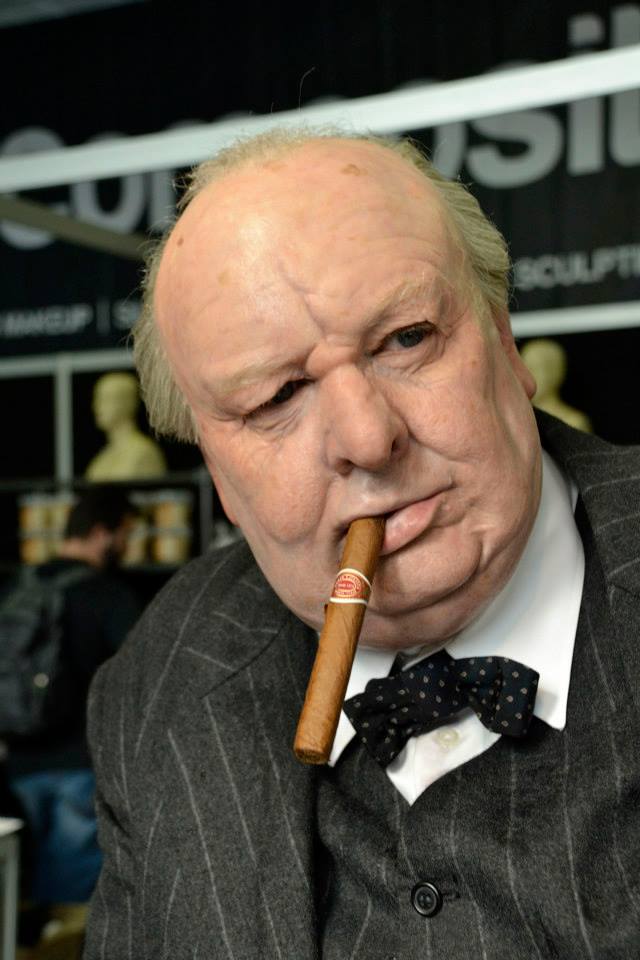 A person dressed as the character Winston Churchill, wearing a pinstripe suit, bow tie, and holding a cigar in their mouth, looks thoughtfully to the side at an indoor event.
