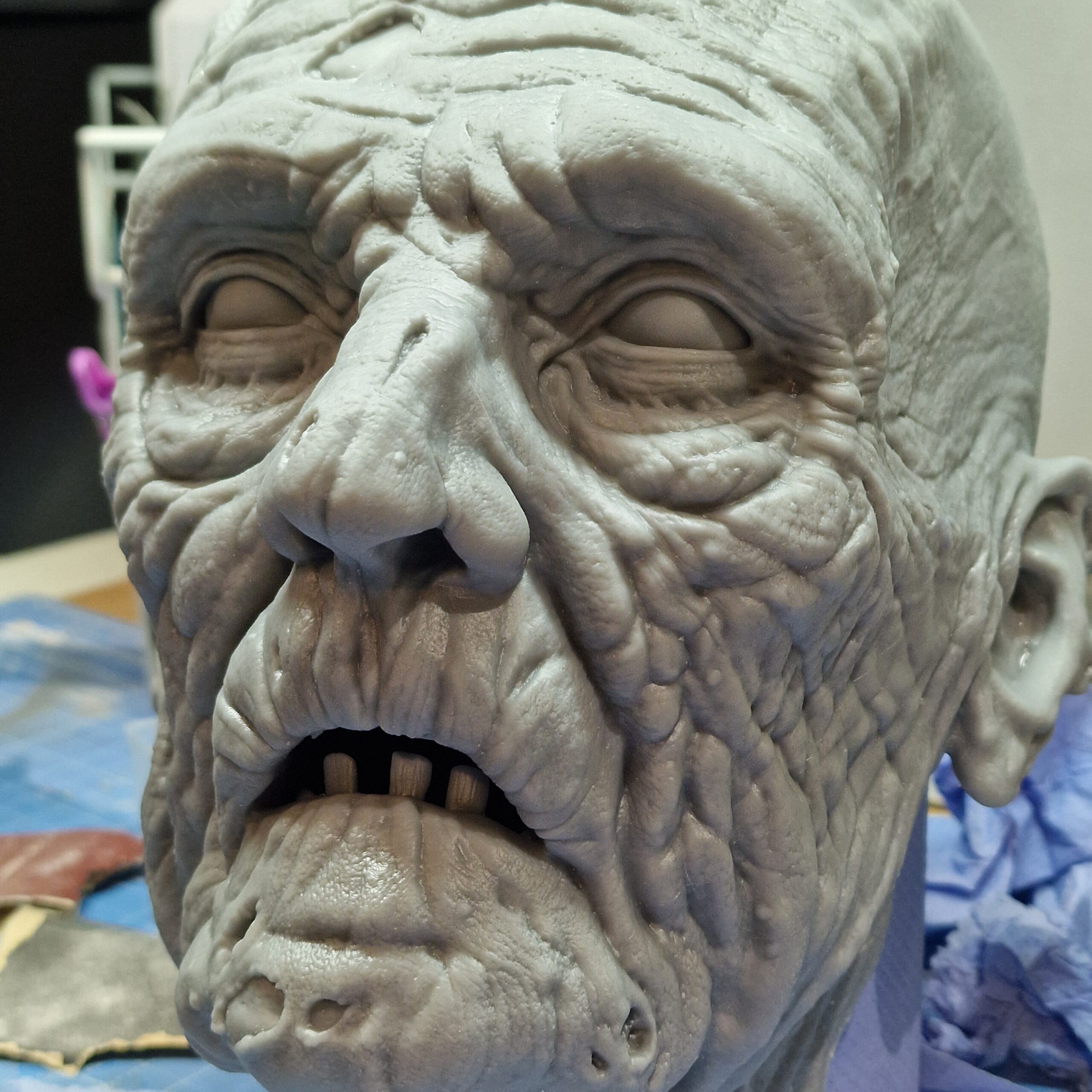 A detailed, realistic sculpture of an elderly mans face with deep wrinkles and textured skin, crafted in gray material using 3D scanning and 3D printing. The eyes are featureless, giving it a lifeless, eerie appearance.