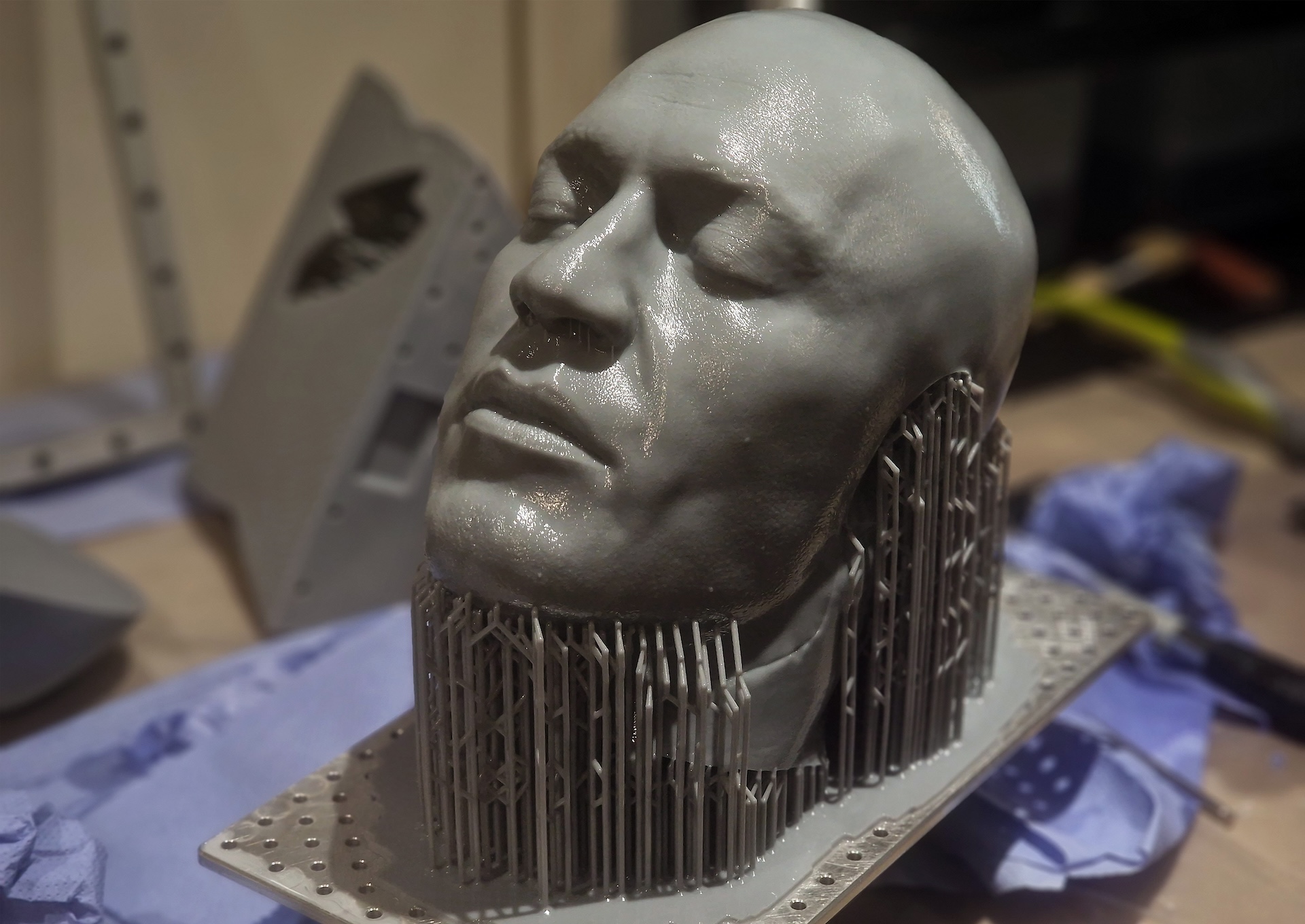 A 3D-printed gray sculpture of a mans head, created using 3d scanning and 3D printing, with closed eyes and intricate support structures, rests on a workspace with blue paper and tools in the background.