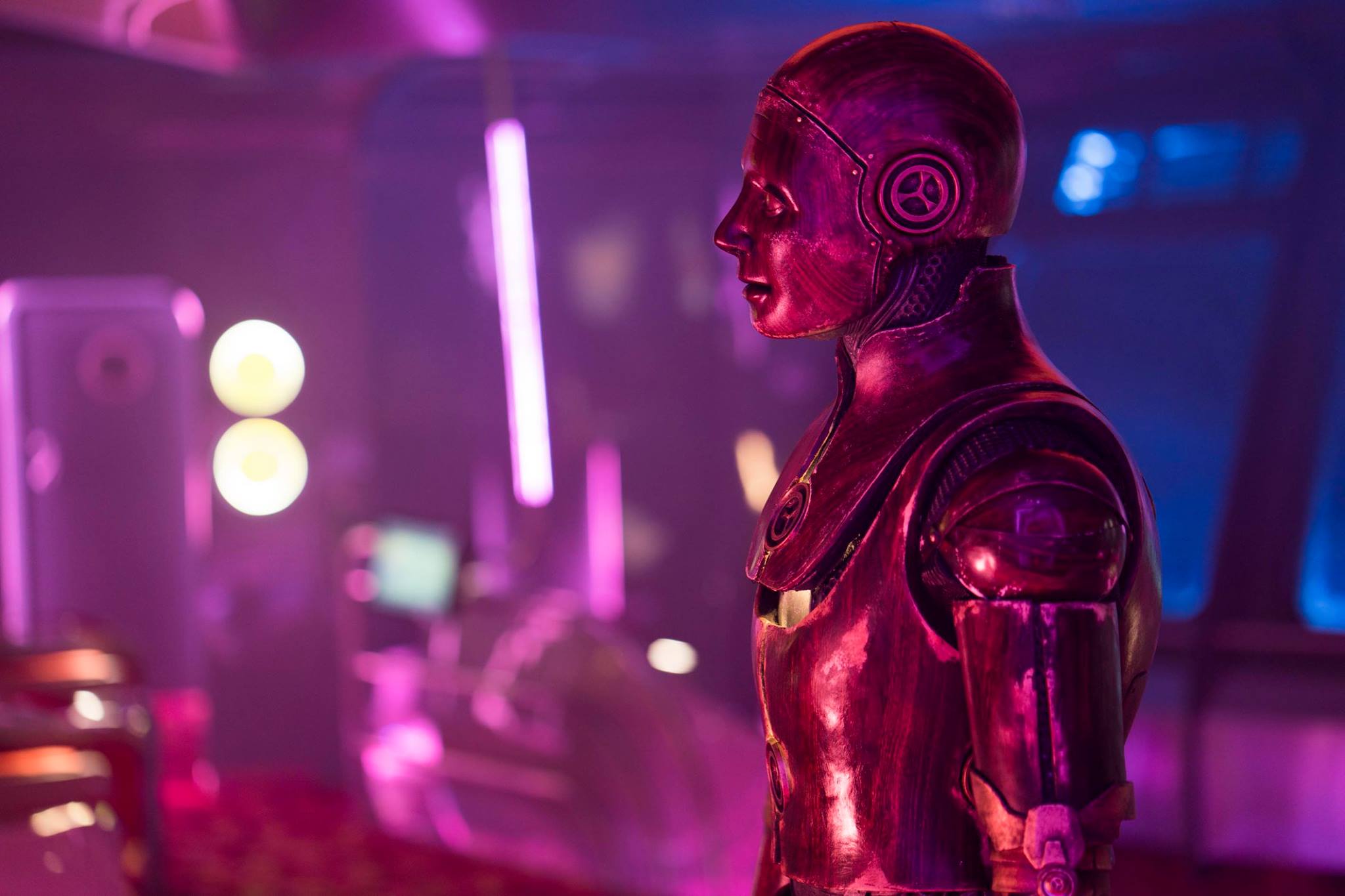 A shiny humanoid robot with a metallic pink-purple finish, crafted with Creature FX artistry, stands in a futuristic, neon-lit room with blurred lights and technology in the background.