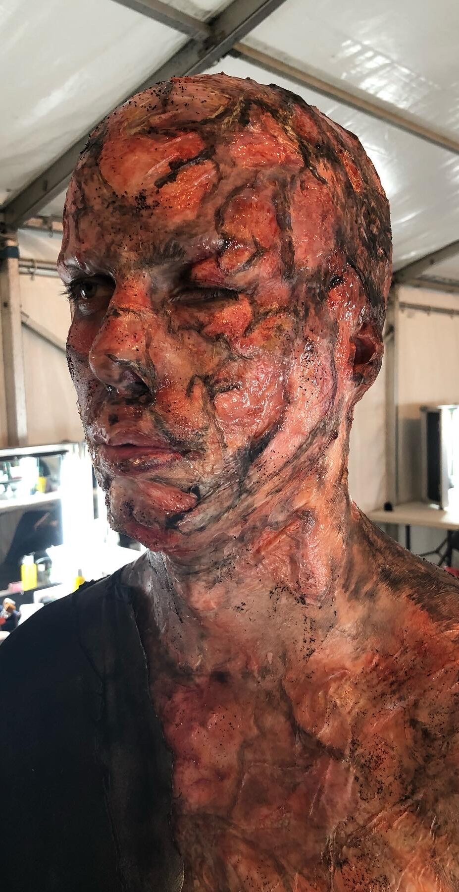 A person with detailed, realistic trauma makeup simulating severe burns and scarring across their face, head, and neck, stands in a well-lit tent or medical workshop.