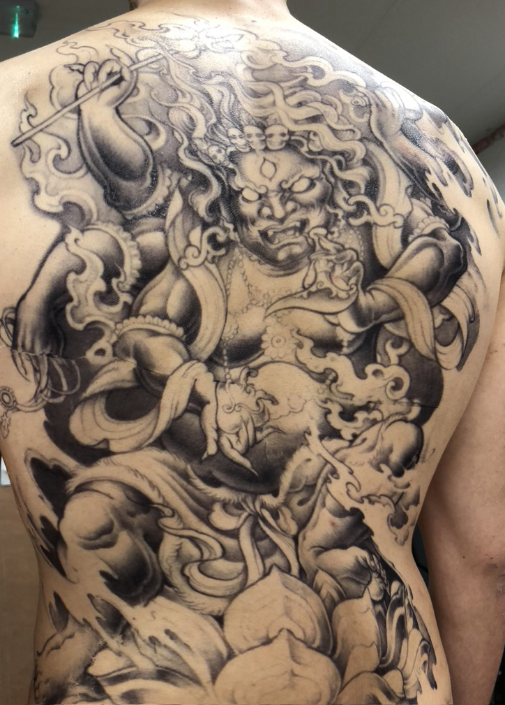 A detailed black and gray tattoo transfer covers a persons back, depicting a fierce, mythological warrior with wild hair, an expressive face, dynamic flowing robes, swirling smoke, and intricate patterns.