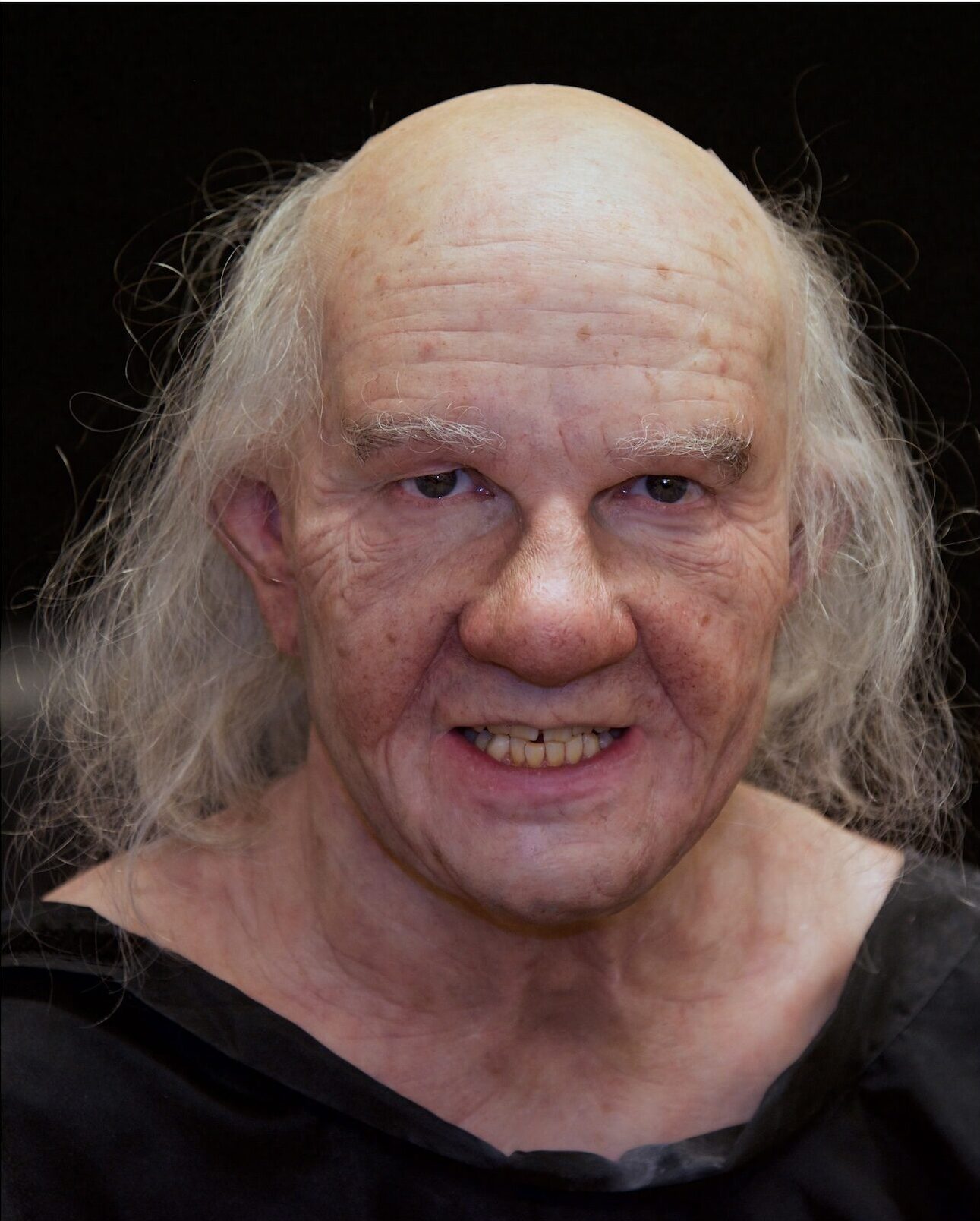 A character with a bald head, long wispy gray hair, aged wrinkled skin, and a slight grimace, wearing a dark garment, is shown against a dark background.