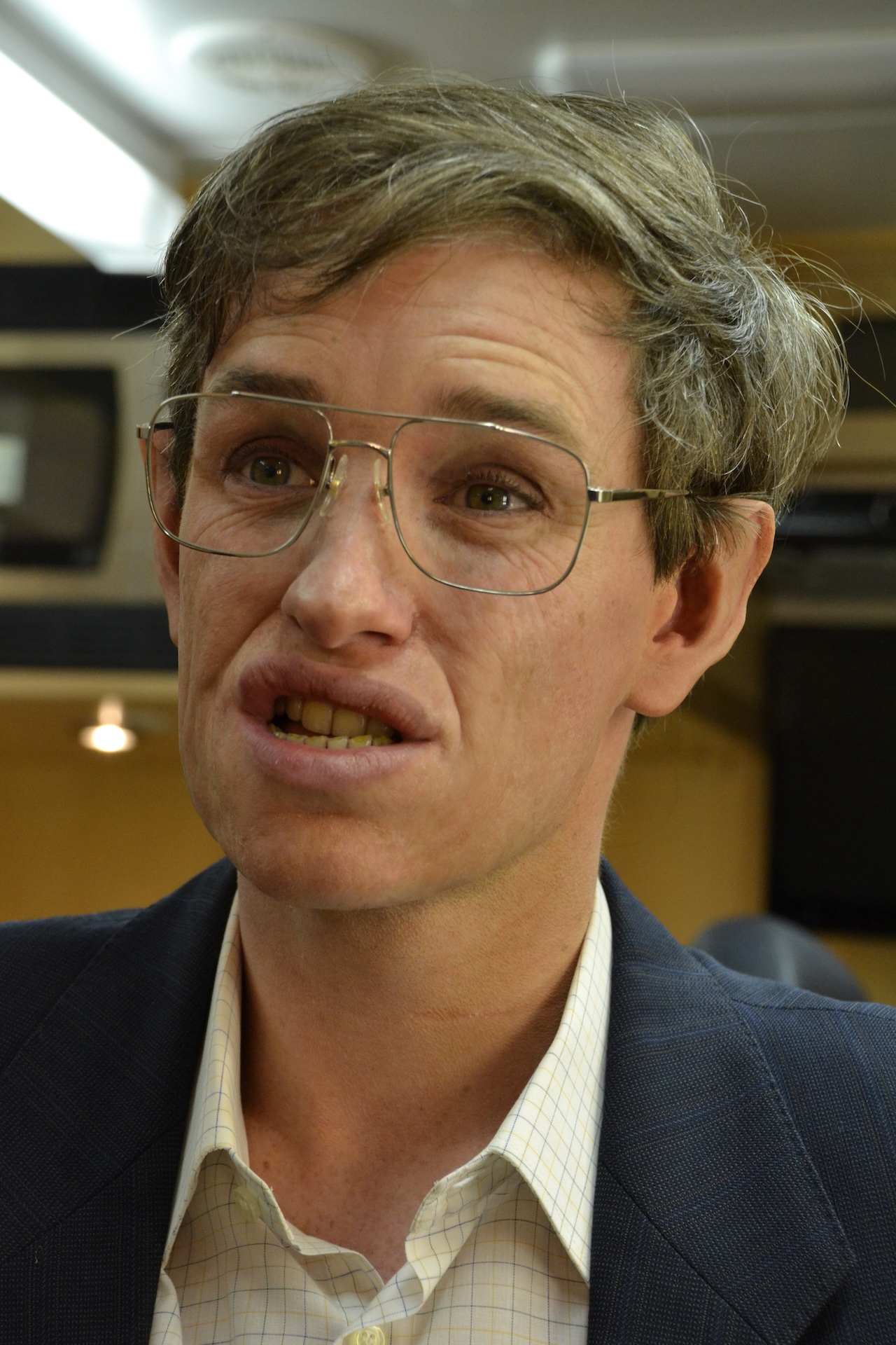 A character with tousled brown hair, wearing glasses, a checked shirt, and a navy blazer appears to be mid-sentence, standing indoors with warm lighting in the background.