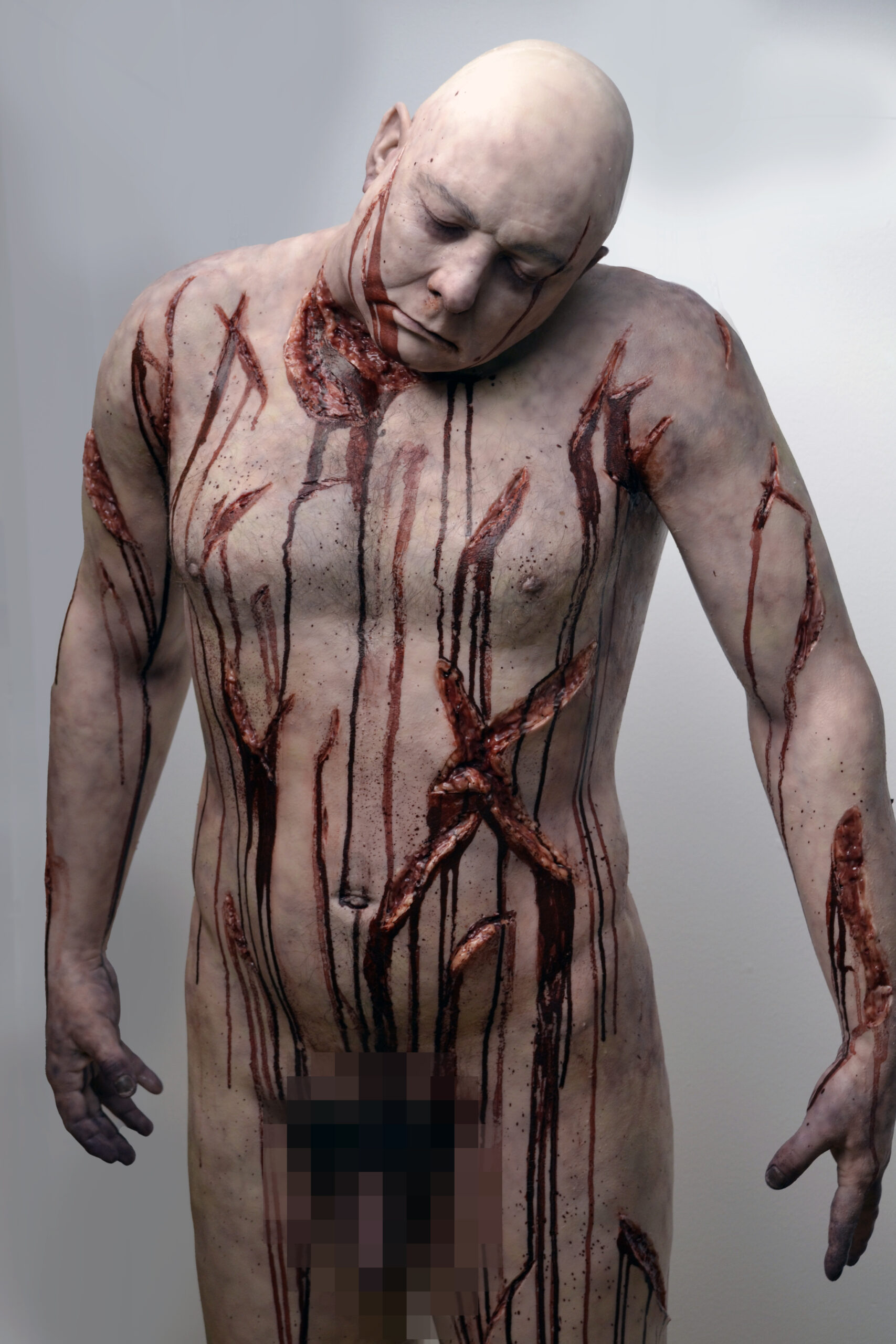 A bald, pale mannequin or figure from Bodies with eyes closed, covered in realistic, deep, bloody wounds and scars on the chest, arms, and torso stands against a plain background. Genital area is pixelated for censorship.