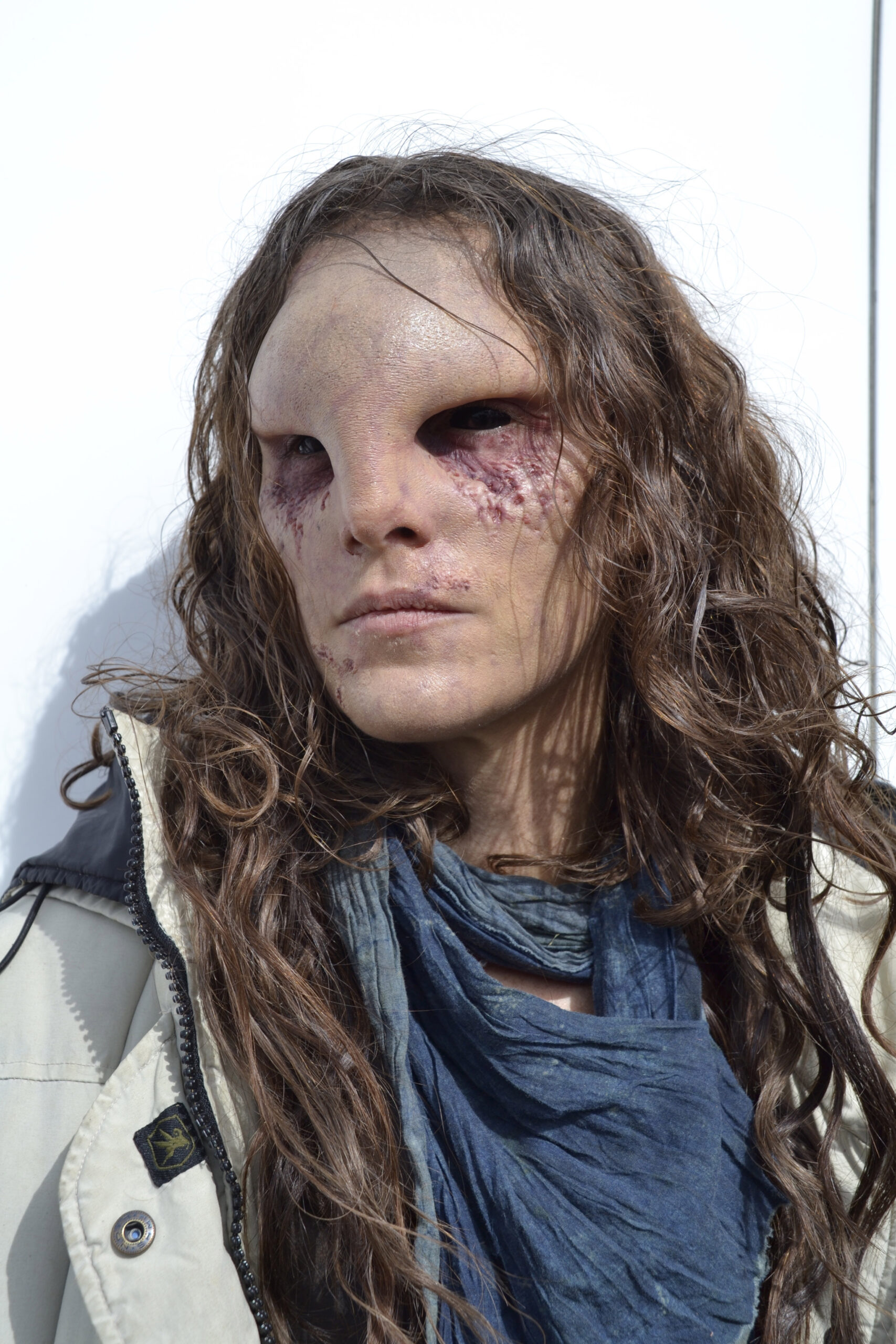 A person with long wavy brown hair and Creature FX facial prosthetics creating an alien-like appearance—large, dark eye sockets and textured skin—wears a cream jacket and layered blue scarf, standing against a white background.