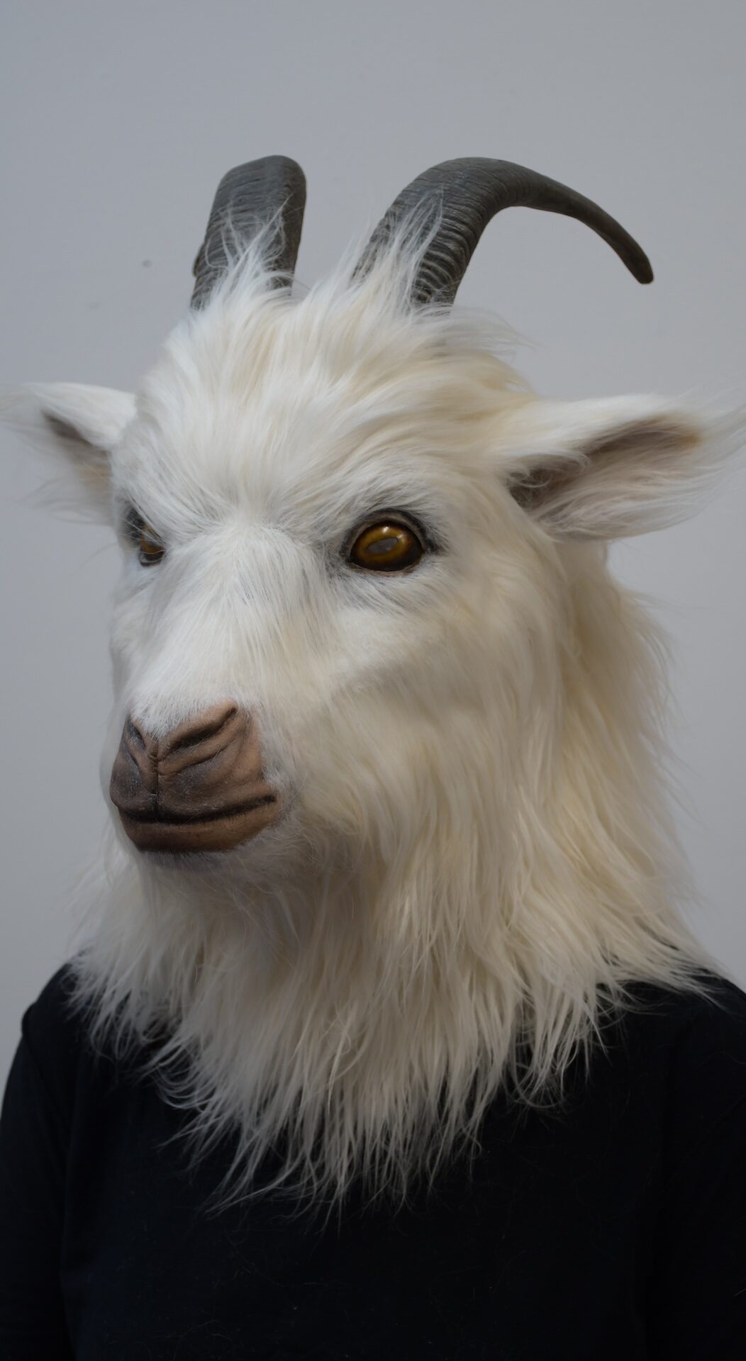 A person wearing a realistic white goat mask with brown horns, furry texture, and golden eyes—crafted with Creature FX expertise—stands against a plain light gray background.