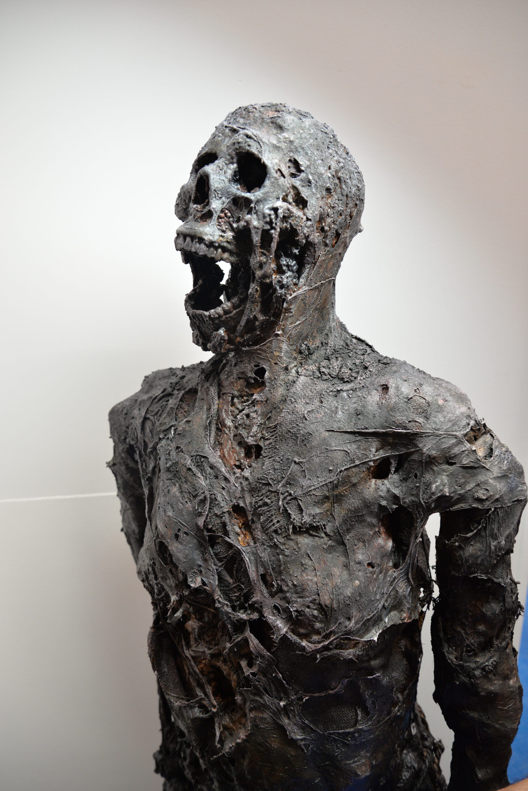 A dark, decayed humanoid sculpture with open mouth and hollow eyes, appearing distressed or in agony, evokes lost bodies with its rough, textured surface and tattered, eroded features.