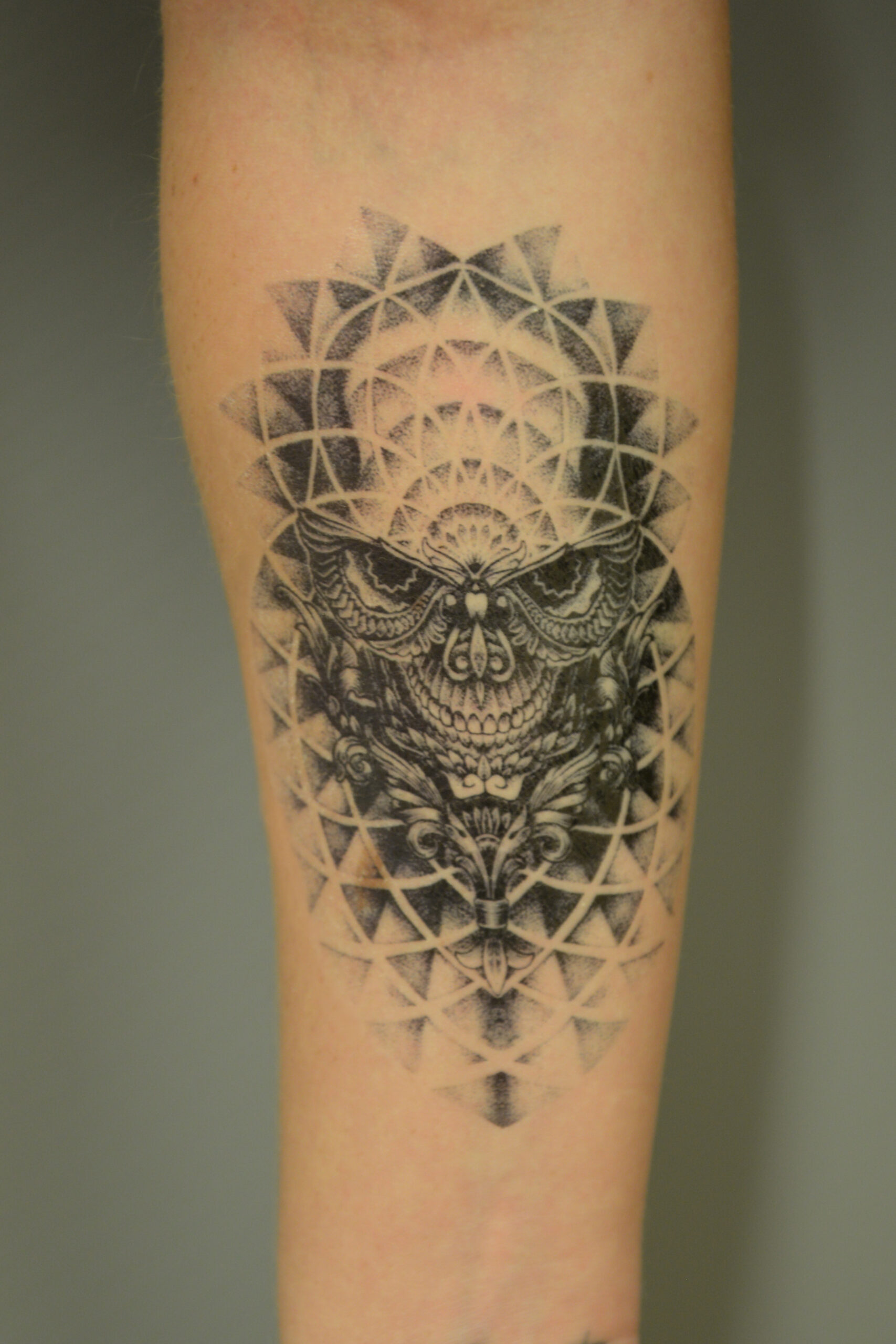 A tattoo transfer of a black ink owl face adorns the forearm, featuring intricate geometric and mandala-like patterns that create a circular, symmetrical design around it.