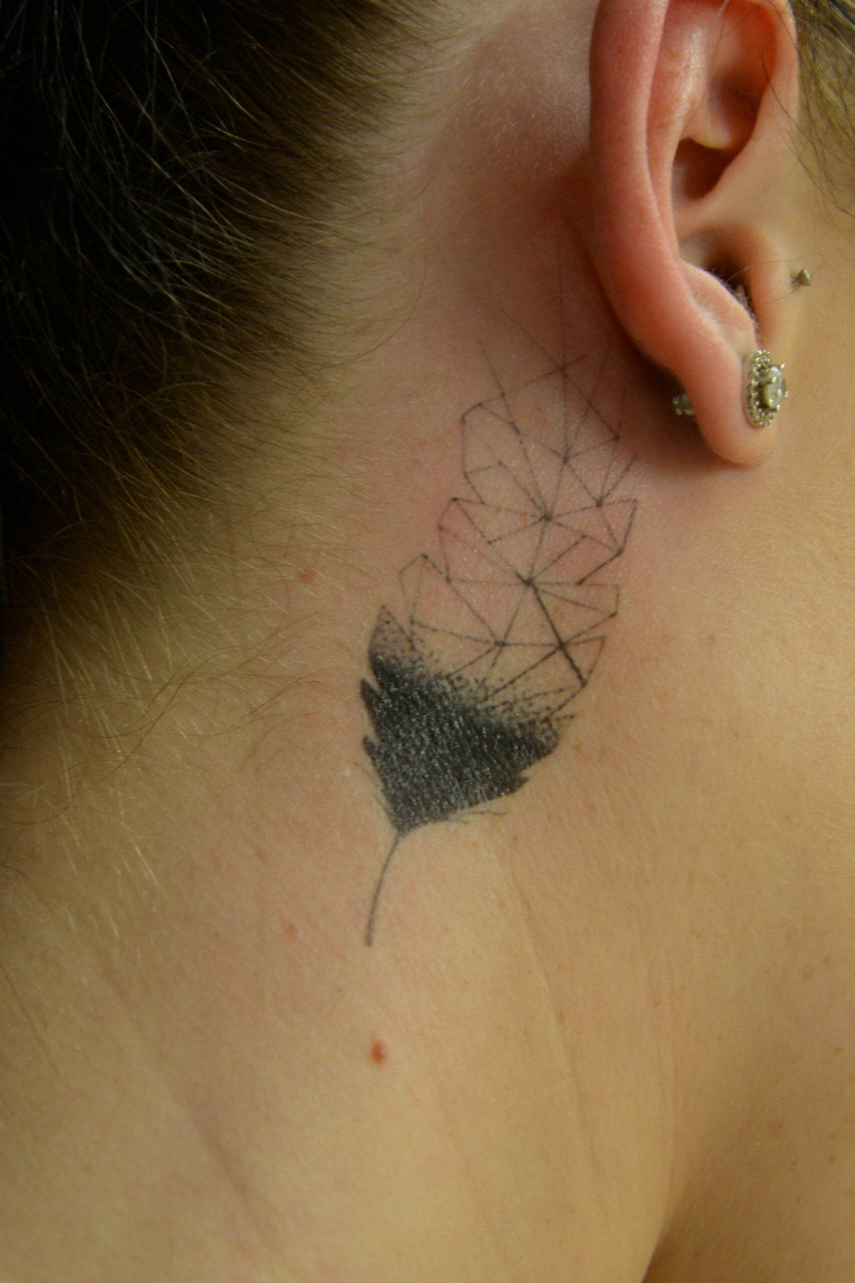 A tattoo transfer behind a person’s ear shows a geometric leaf design, with the lower half shaded and the upper half outlined in angular shapes. The person has light skin and is wearing a small decorative earring.