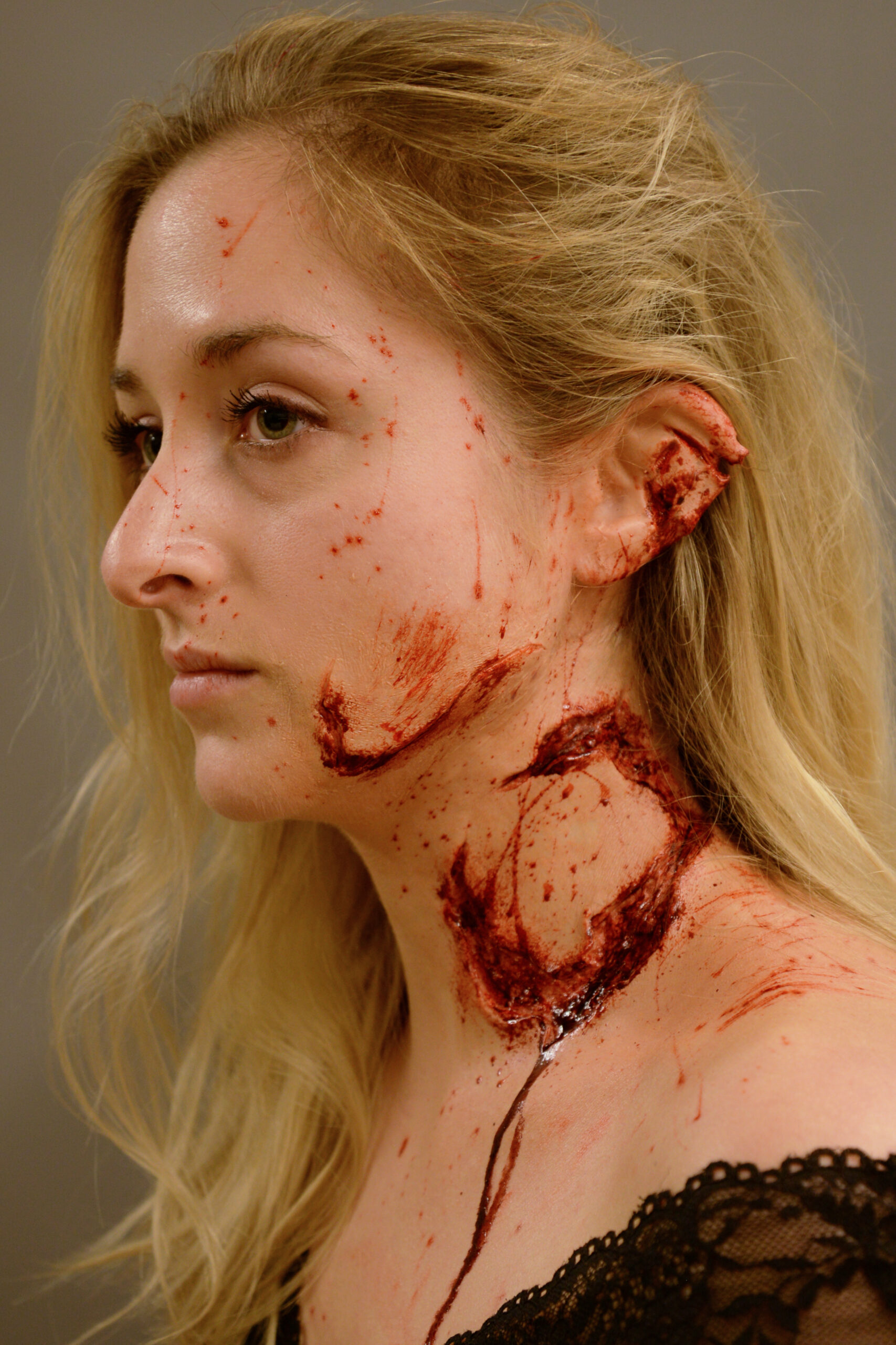 A woman with long blonde hair has realistic-looking trauma makeup on her face and neck, showing fake blood, wounds, and a partially torn ear, creating a dramatic and gruesome appearance against a plain background.