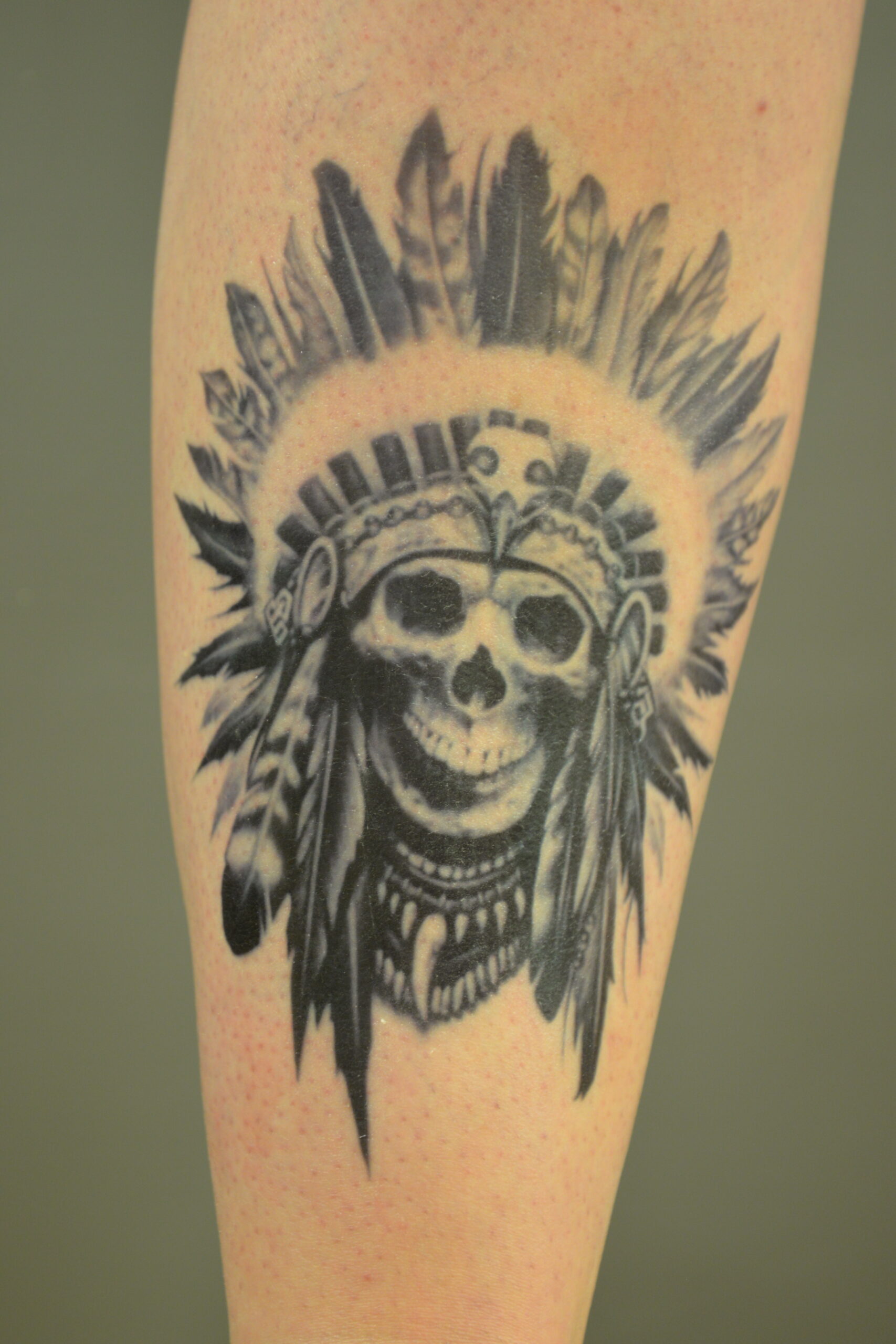 A black and gray Tattoo Transfer of a skull wearing a feathered Native American headdress, intricately detailed with beads and feathers, inked on a persons skin.
