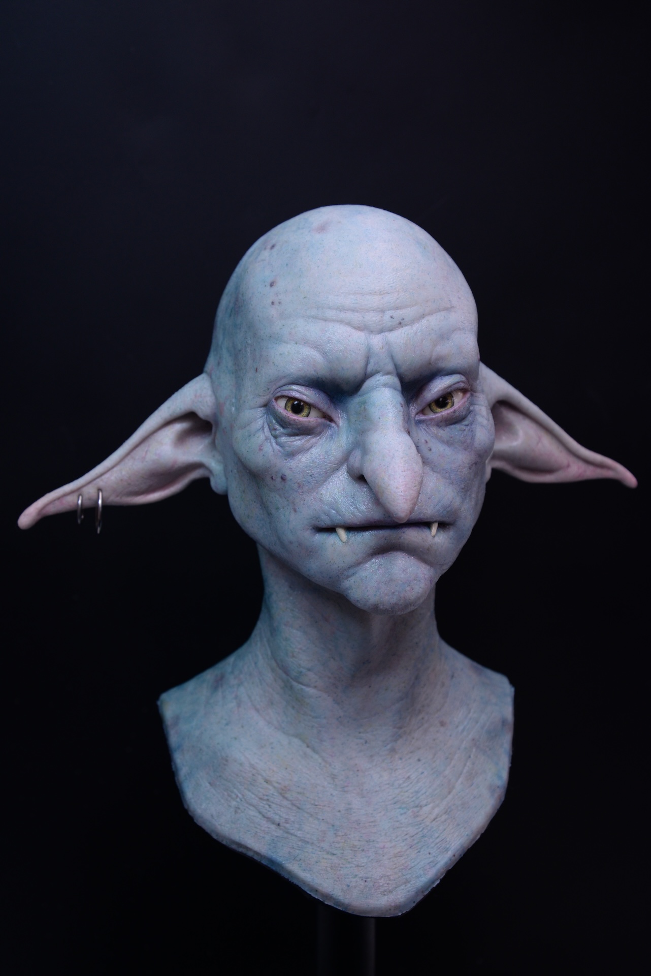 A realistic VFX reference sculpture of a blue-skinned goblin with pointed ears, large nose, small fangs, and a serious expression, set against a black background. One ear features two silver hoop earrings.