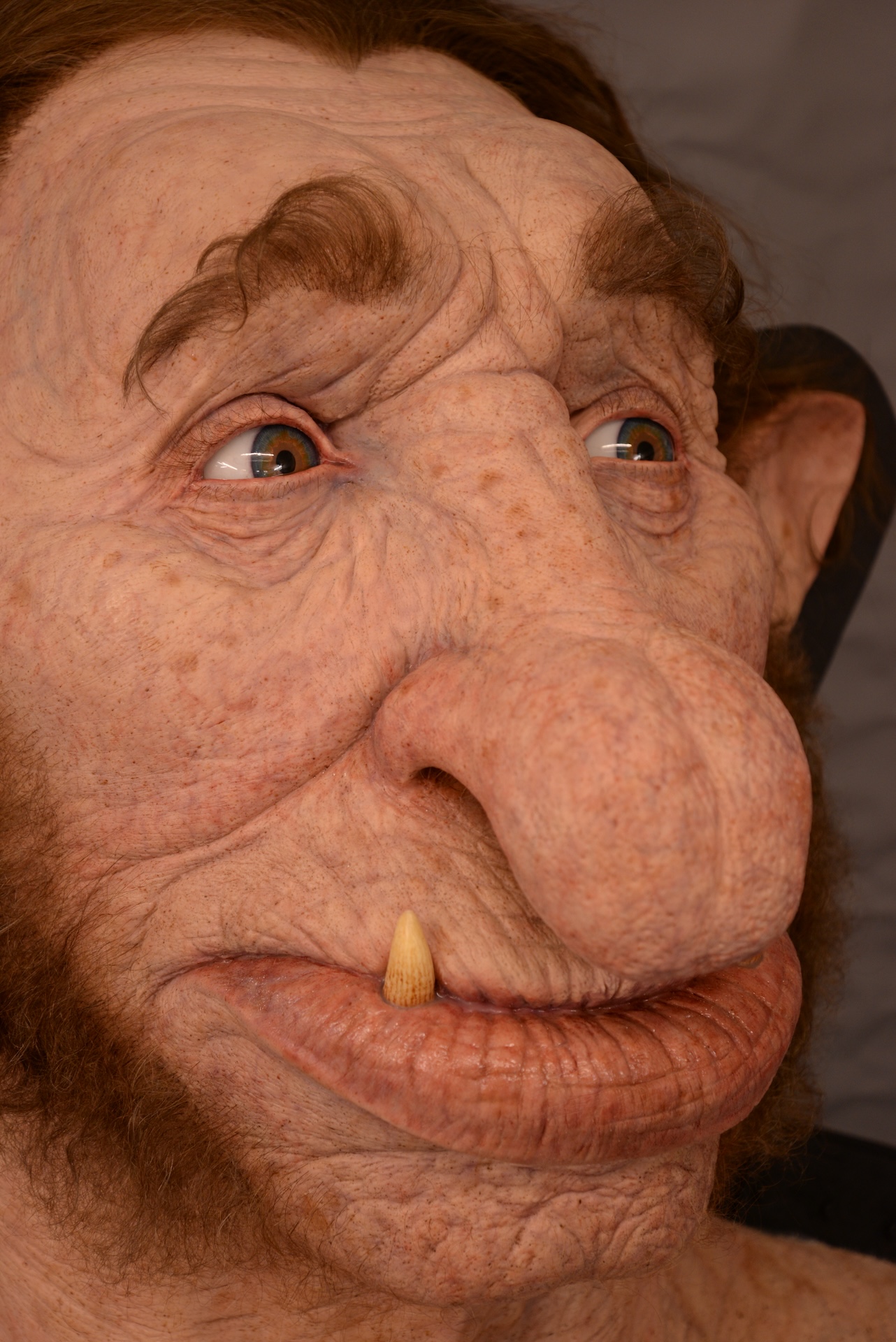 A close-up of a detailed troll or ogre mask with exaggerated features, including a large nose, bushy eyebrows, one protruding tooth, and textured, pinkish skin—ideal as a VFX reference for creature design.