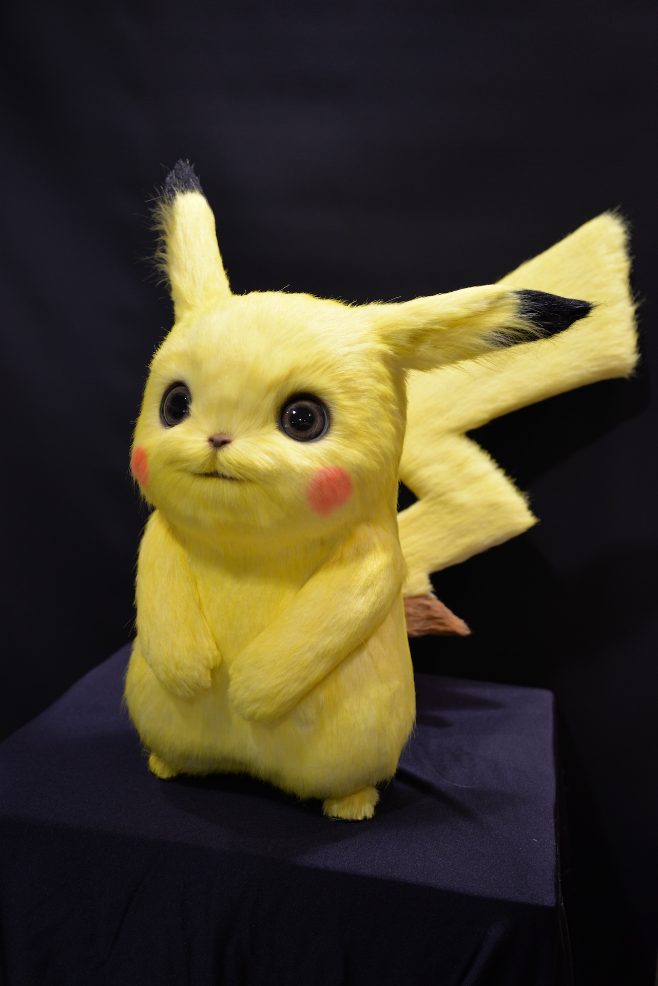 A realistic, fluffy Pikachu model with bright yellow fur, large black eyes, red cheeks, and pointed ears stands on a dark fabric-covered surface against a black background—perfect as a VFX reference.