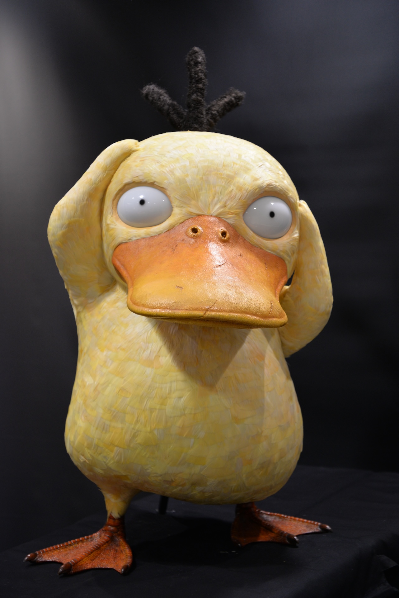 A large, lifelike model of Psyduck, perfect as a VFX reference, stands against a dark background. The yellow duck-like Pokémon clutches its head with both hands, wide eyes and orange beak highlighted under dramatic lighting.