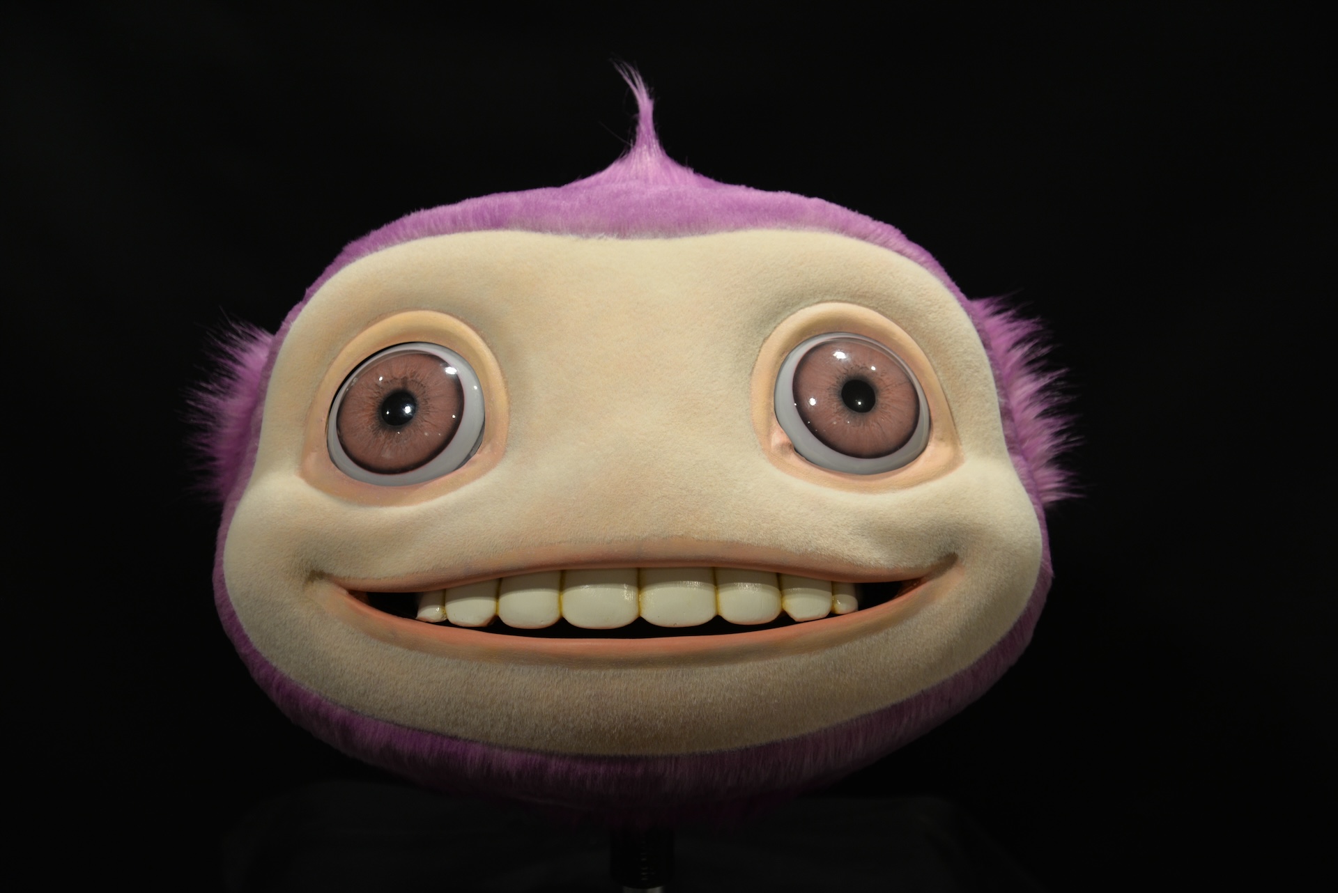 A round, plush creature with purple fur, a cream-colored face, wide eyes, and a big toothy smile sits against a black background—perfect as a VFX reference for animated character design.