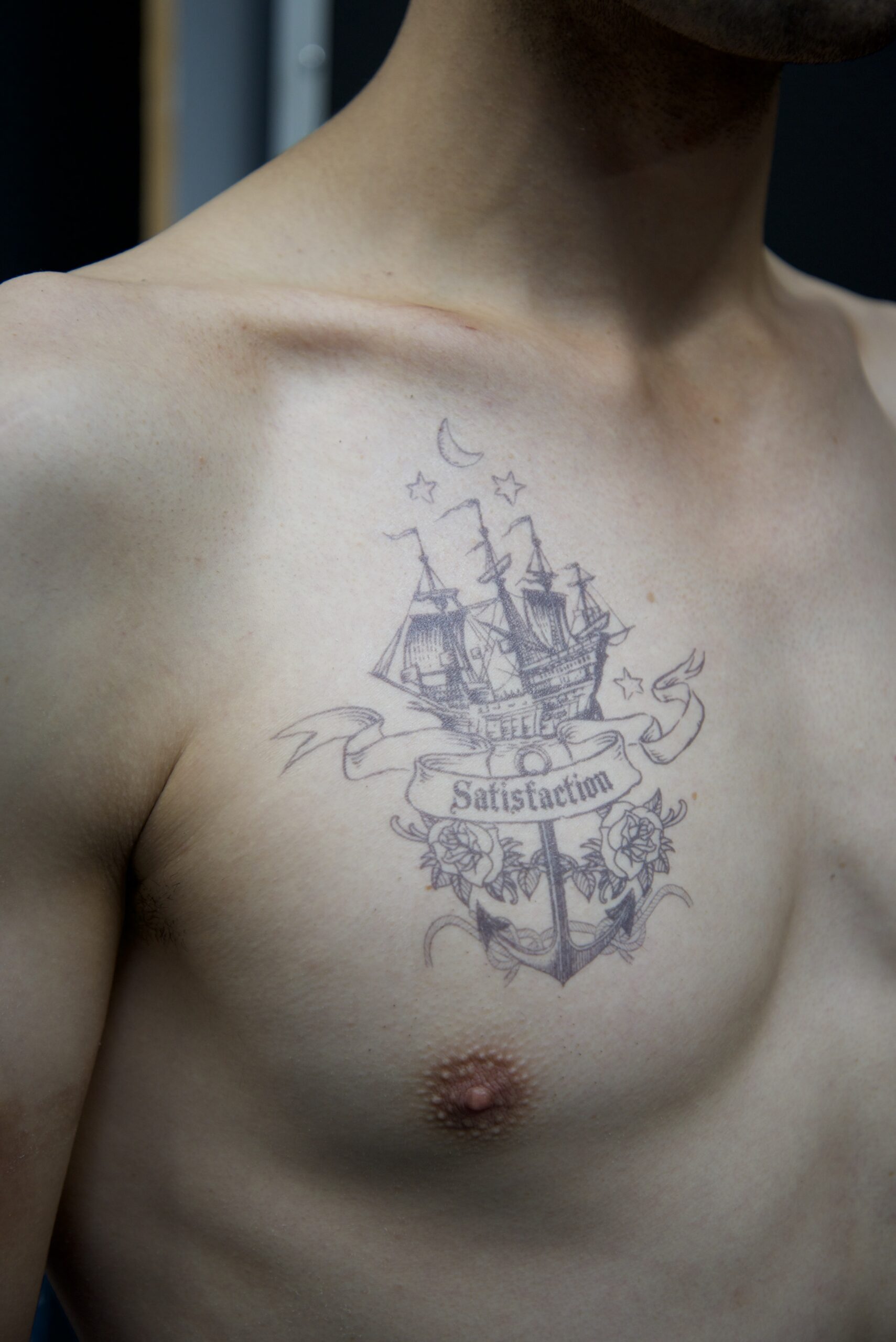 A Tattoo Transfer on a persons chest shows a sailing ship, stars, a crescent moon, roses, and a banner with the word Satisfaction. The detailed design covers the upper left side of the chest.