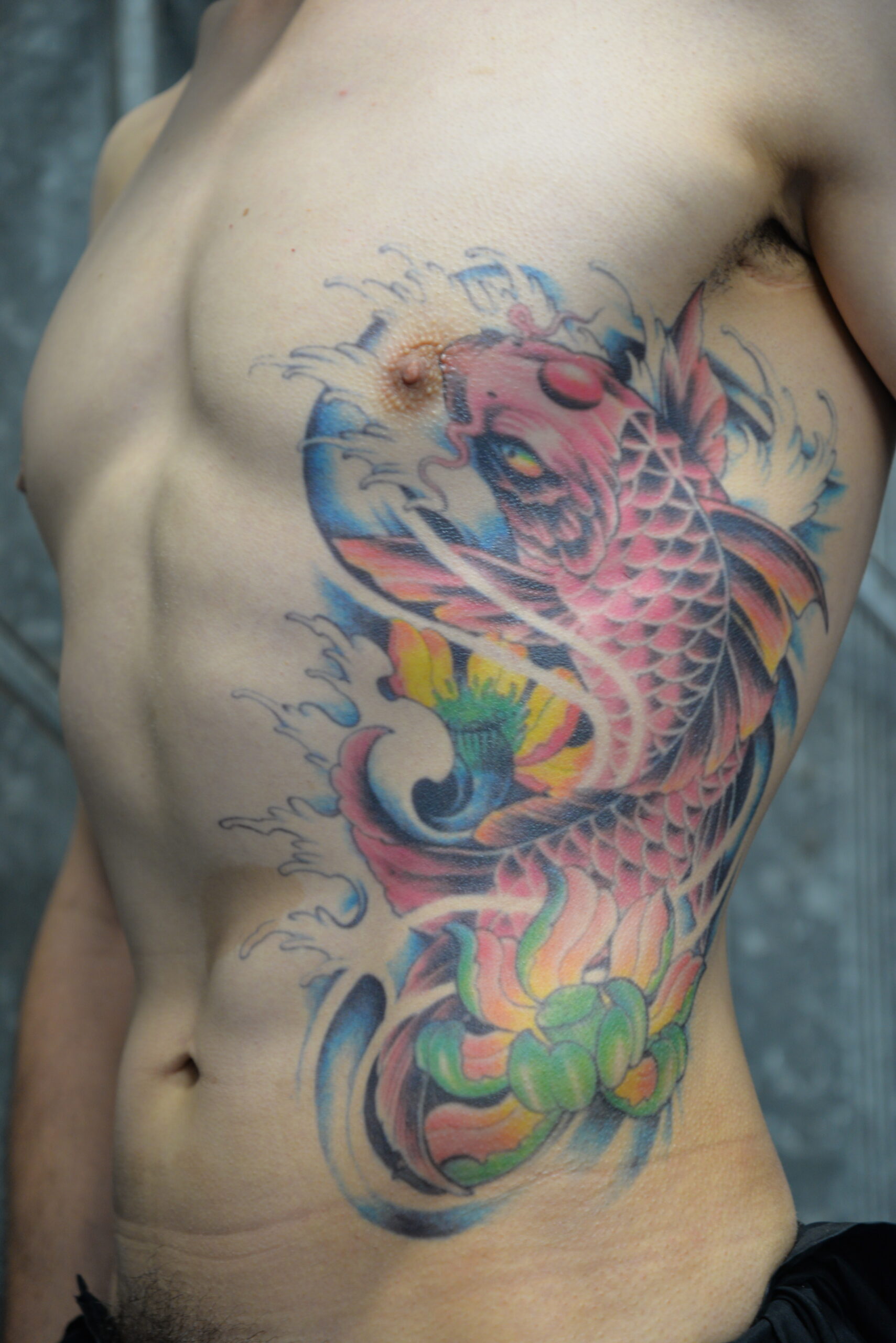 A colorful koi fish tattoo, created with a precise tattoo transfer, features waves and floral elements cascading along the side of a person’s ribcage and torso.