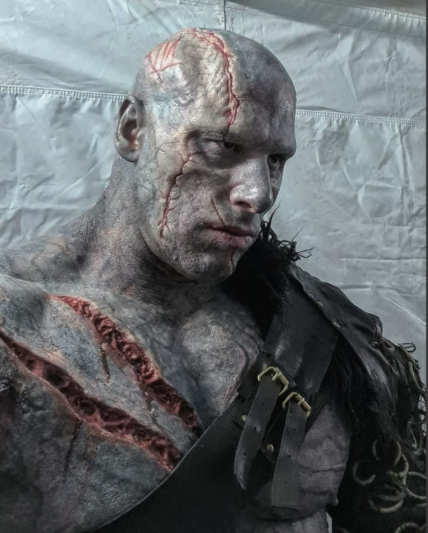 A person in detailed Creature FX monster makeup with grayish skin, prominent scars, and red wounds on the chest, wearing a leather strap costume, stands against a plain white background.
