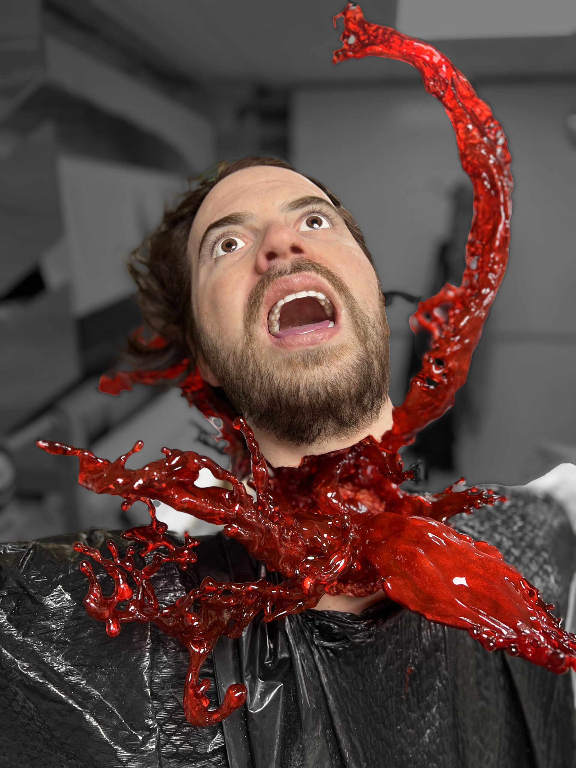 A man with a shocked expression appears to have a vivid, 3D printed, bright red wound on his neck, with liquid splashing out, resembling a dramatic special effects scene. The background is blurred in grayscale.