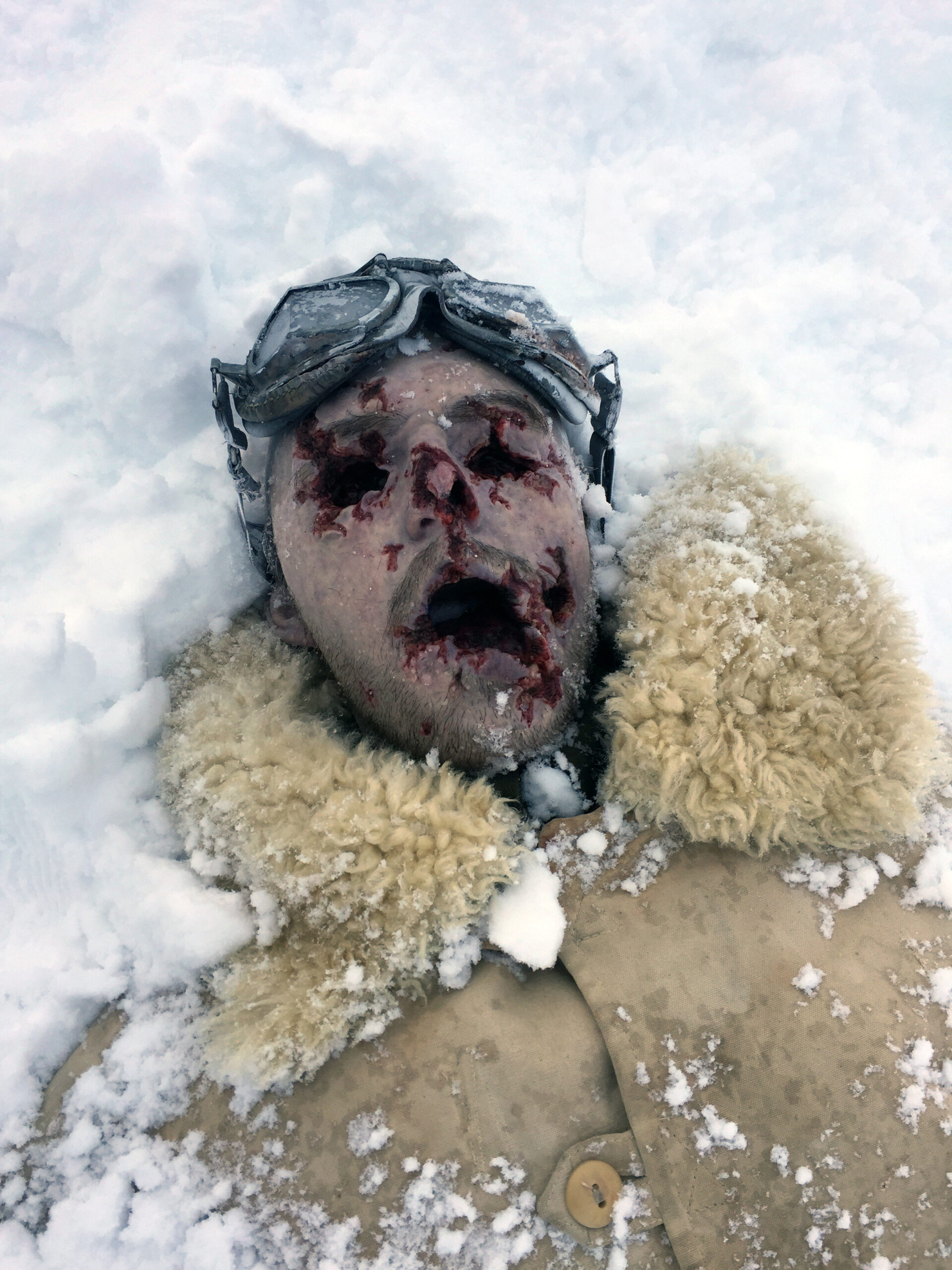 A mannequin with a bloody, damaged face and vintage aviator goggles lies partially buried in the snow, its tan shearling-lined coat evoking memories of lost bodies in forgotten winters.