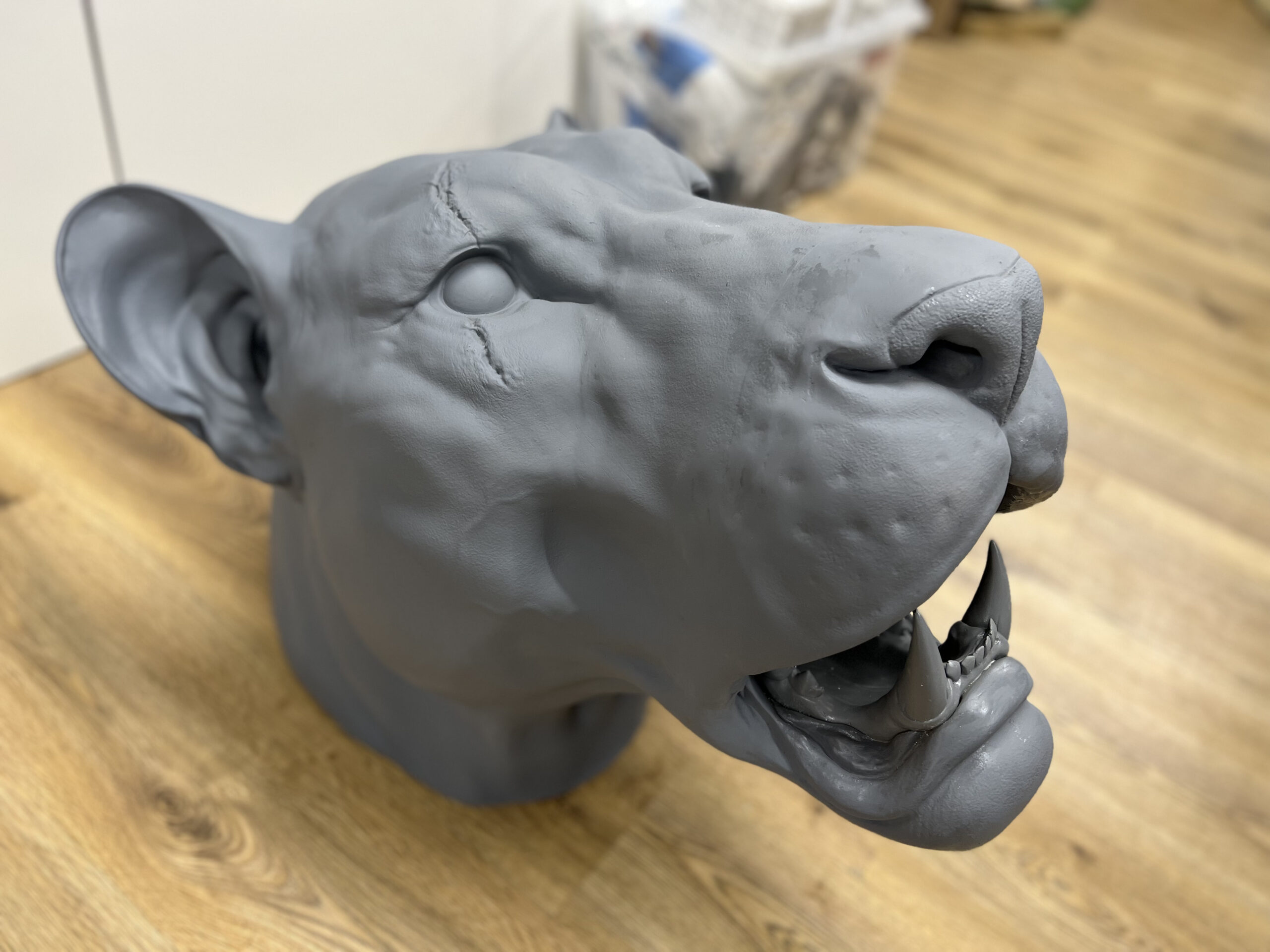 A gray, unfinished lion or big cat head sculpture with detailed features, created using 3D scanning, sits with an open mouth on a wooden floor, surrounded by some out-of-focus household items in the background.