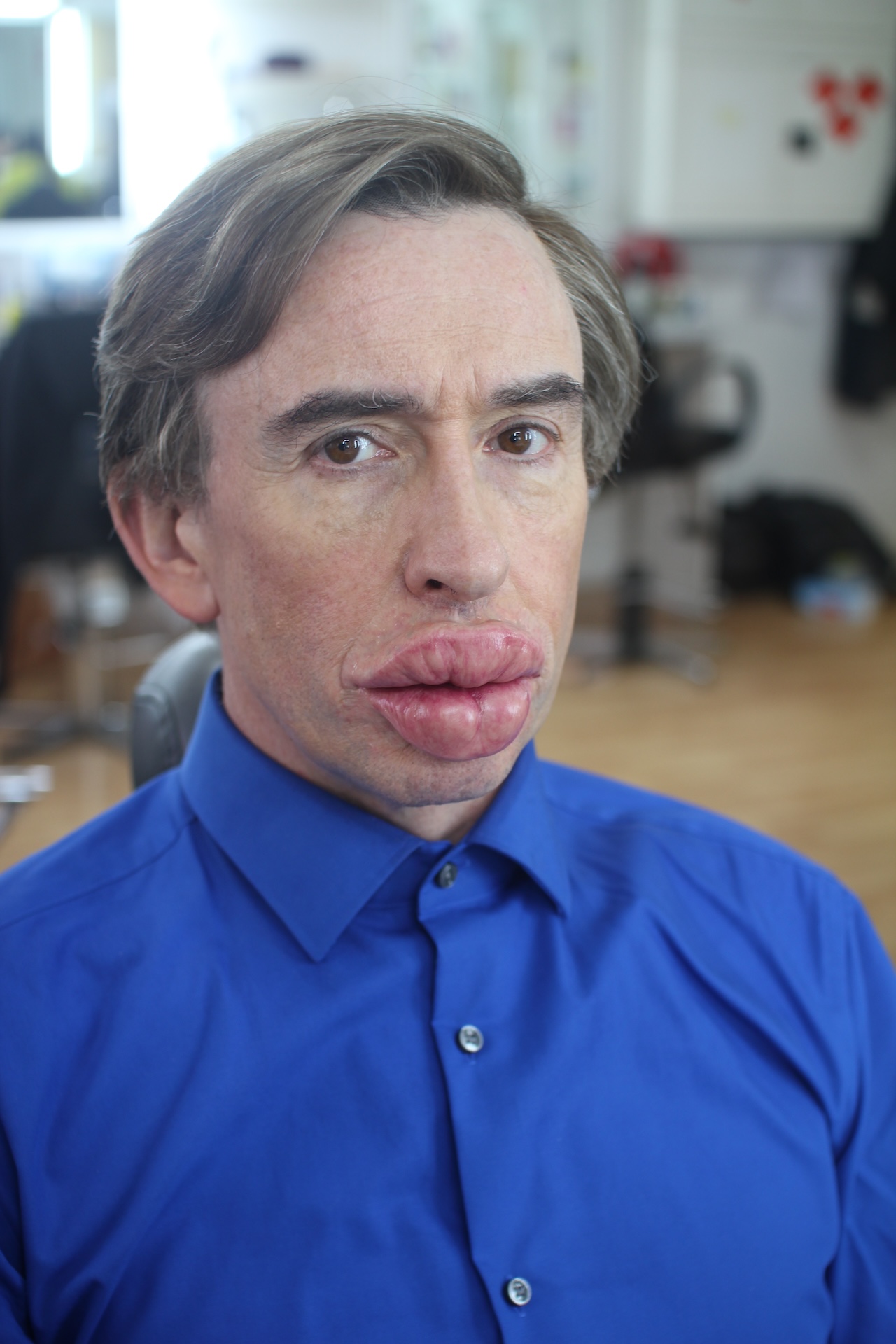 A character with neatly combed hair, wearing a bright blue button-up shirt, sits indoors. His lips appear unusually large and swollen, possibly due to prosthetic makeup. The background shows salon chairs and blurred objects.