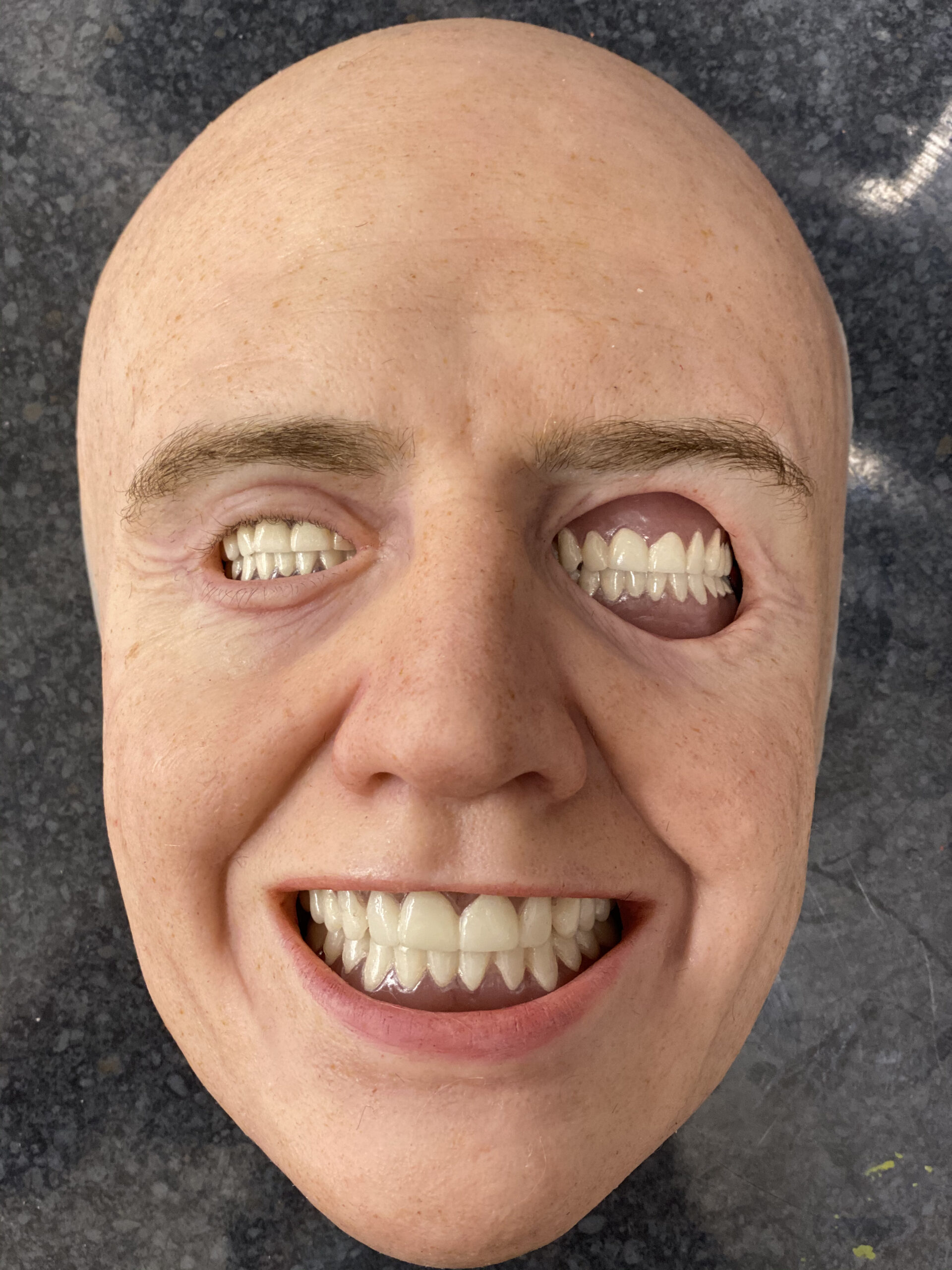 A realistic, bald human mask for VFX reference features an unsettling detail: one eye socket is replaced with a smiling mouth full of teeth. The mask sports a broad grin and rests on a gray speckled surface.