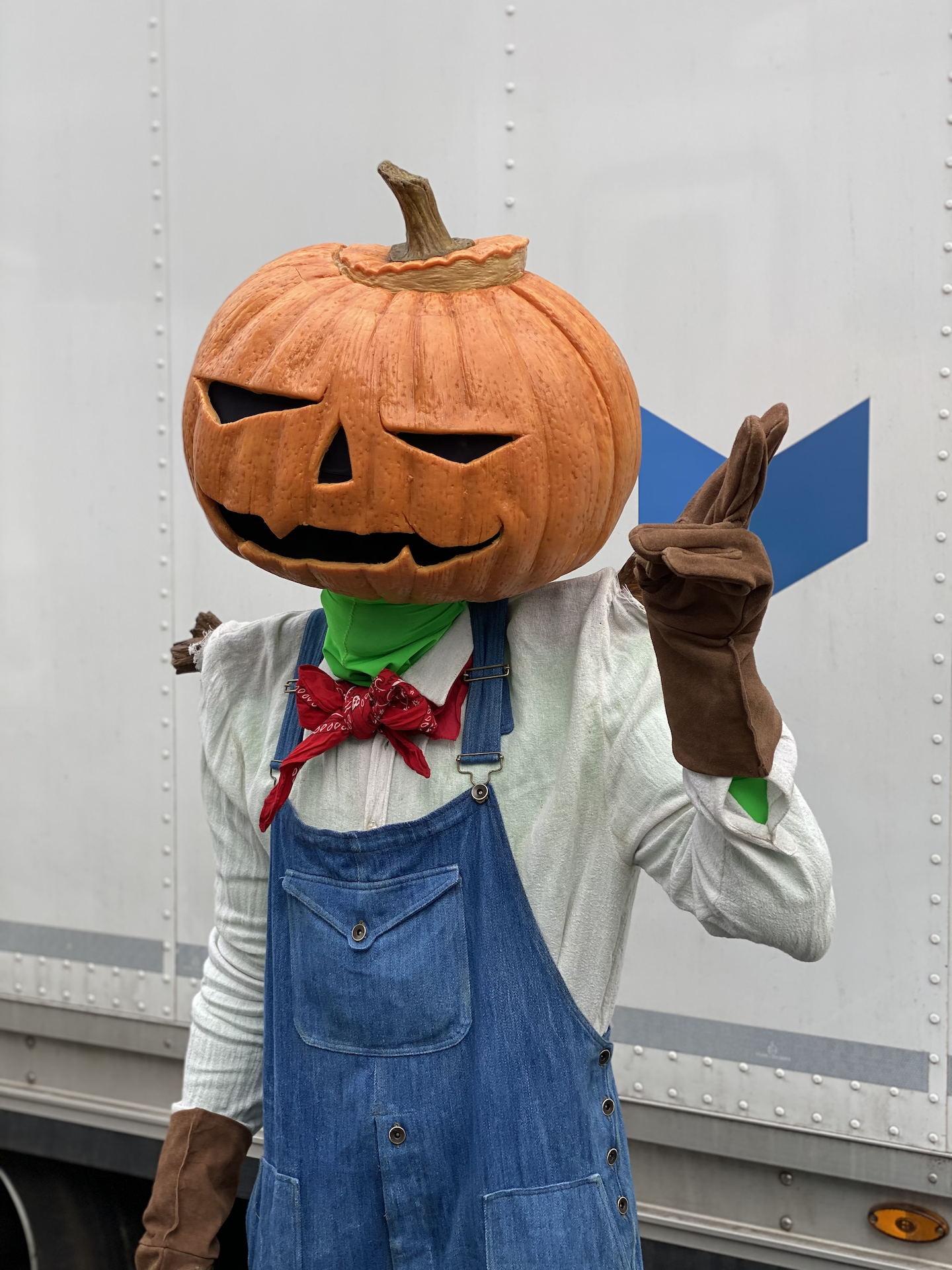 A person wearing a large carved pumpkin mask, crafted with Creature FX artistry, denim overalls, brown gloves, a red bandana, and a white shirt poses with one hand raised against a plain background.