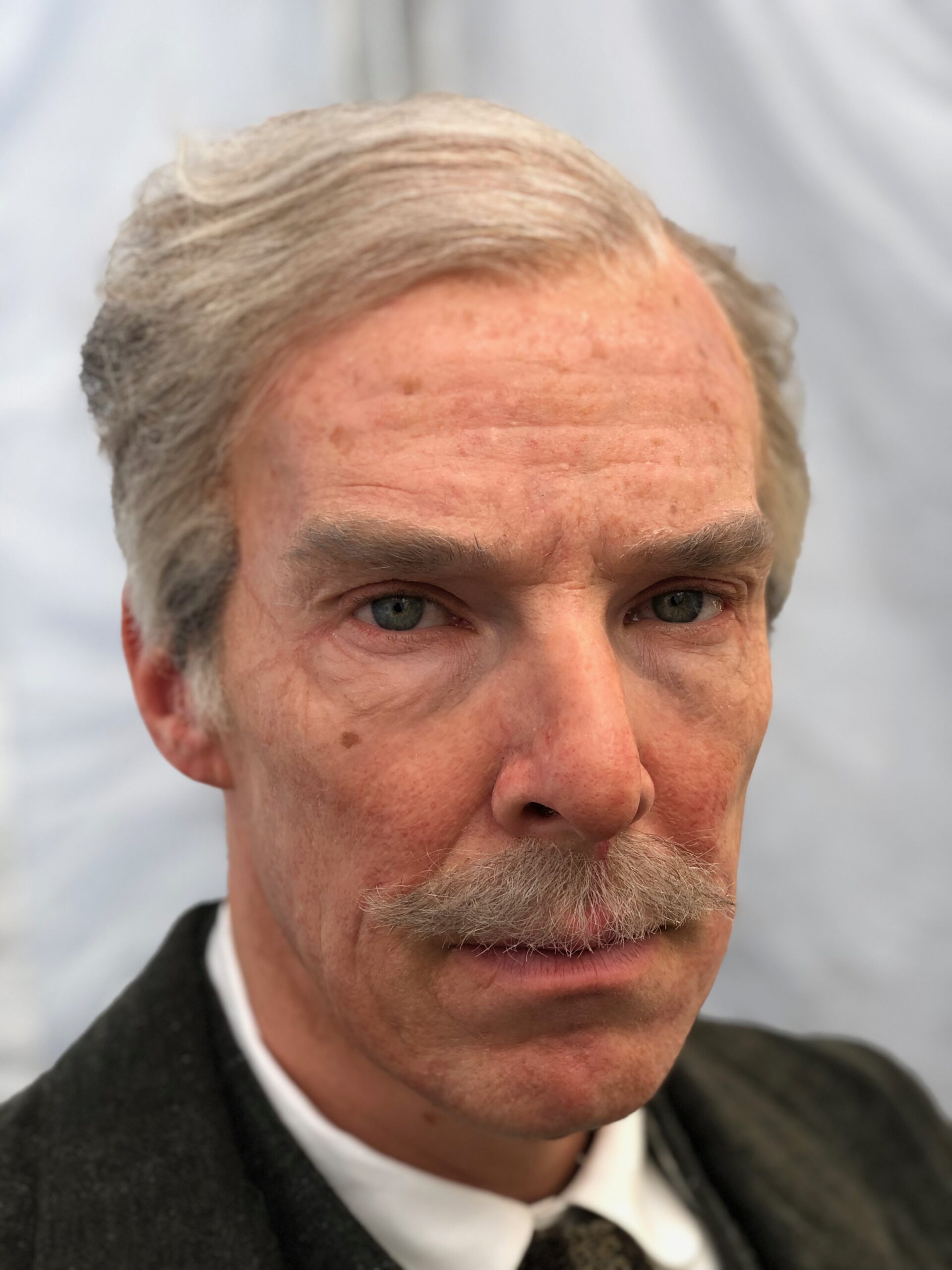 A distinguished character with light gray hair and a mustache, dressed in a dark suit and tie, stares seriously at the camera. The background features softly blurred, light-colored fabric.