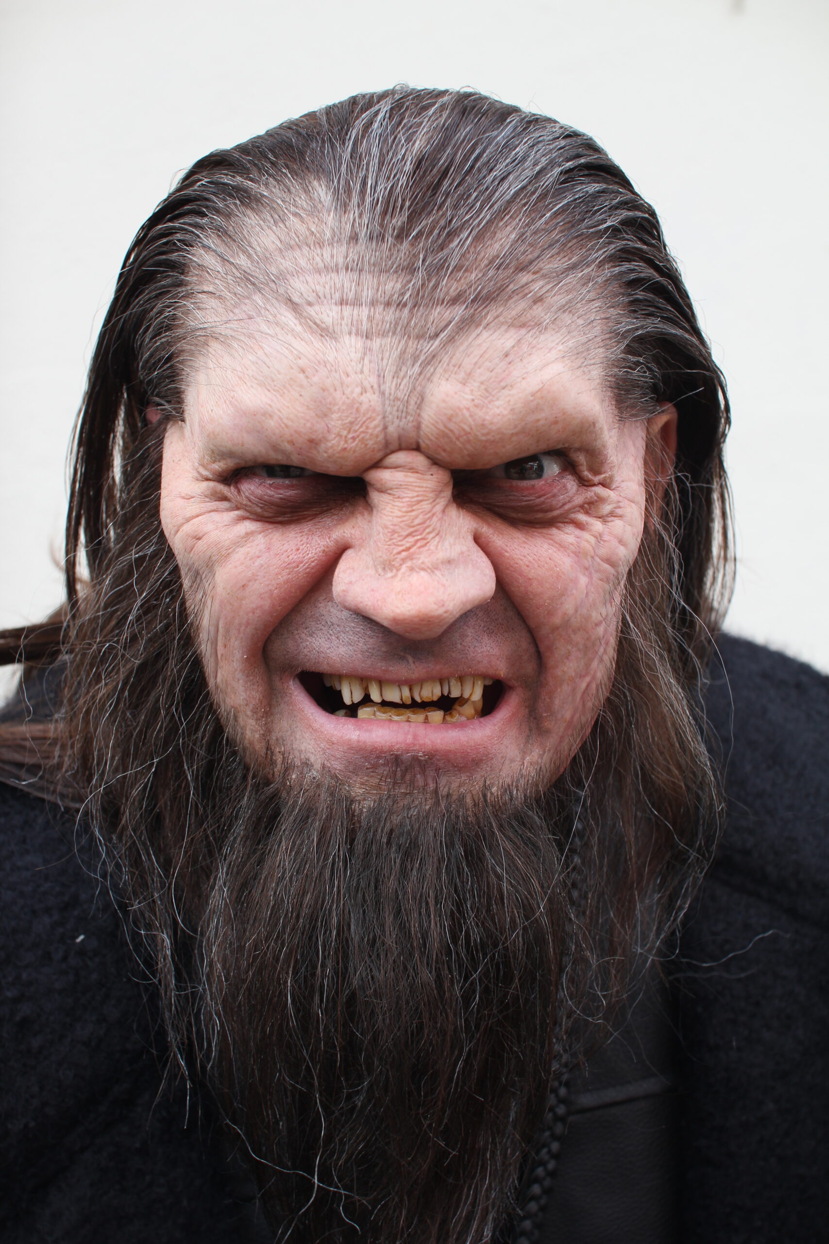 A person wearing realistic Creature FX werewolf makeup, with long dark hair, a full beard, sharp yellow teeth, and wrinkled skin, snarling at the camera against a plain background.