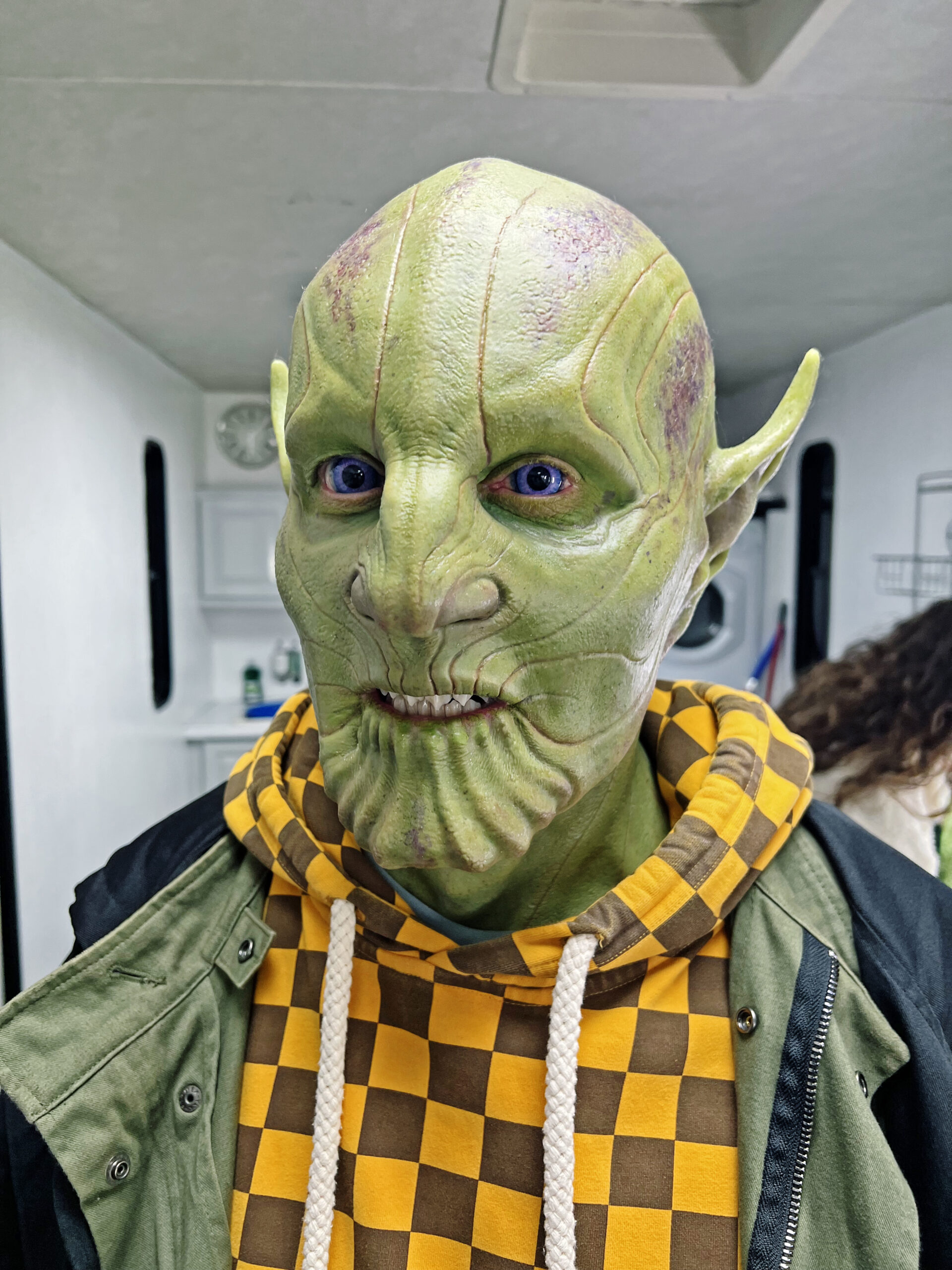 A person wearing detailed green alien Creature FX makeup with pointed ears and ridged skin, dressed in a yellow and brown checkered hoodie and a green jacket, standing indoors.