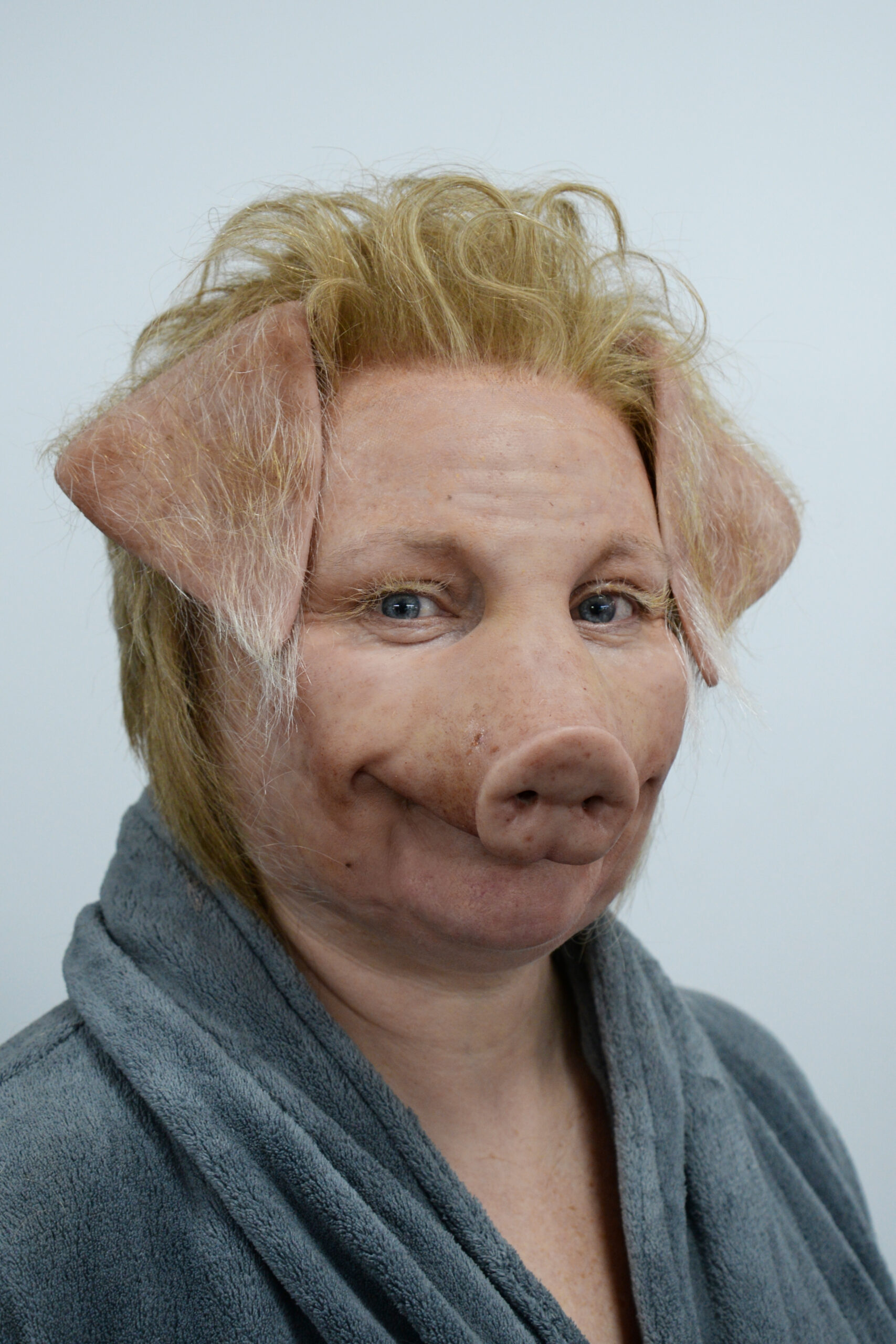 A person wearing a pig mask with realistic Creature FX features, including a pig snout and ears, and a blond wig, dressed in a gray robe against a plain light background.