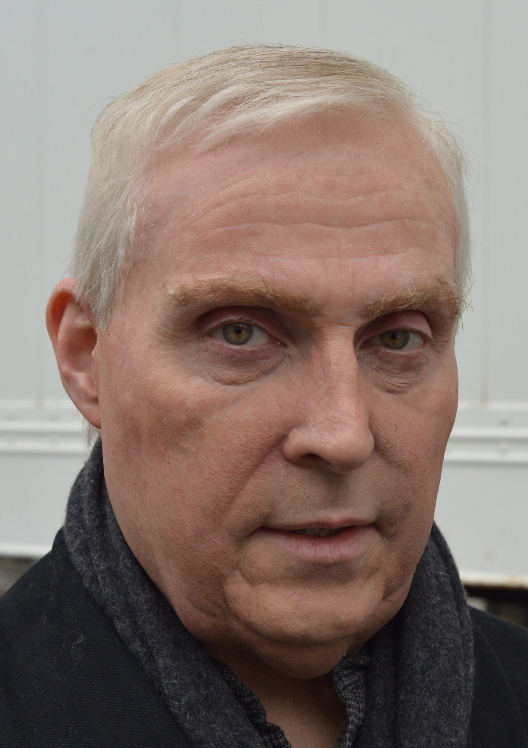A middle-aged man with short, light gray hair and fair skin wears a dark scarf and looks directly at the camera with a neutral expression, standing in front of a light-colored background that subtly highlights KM Effects.