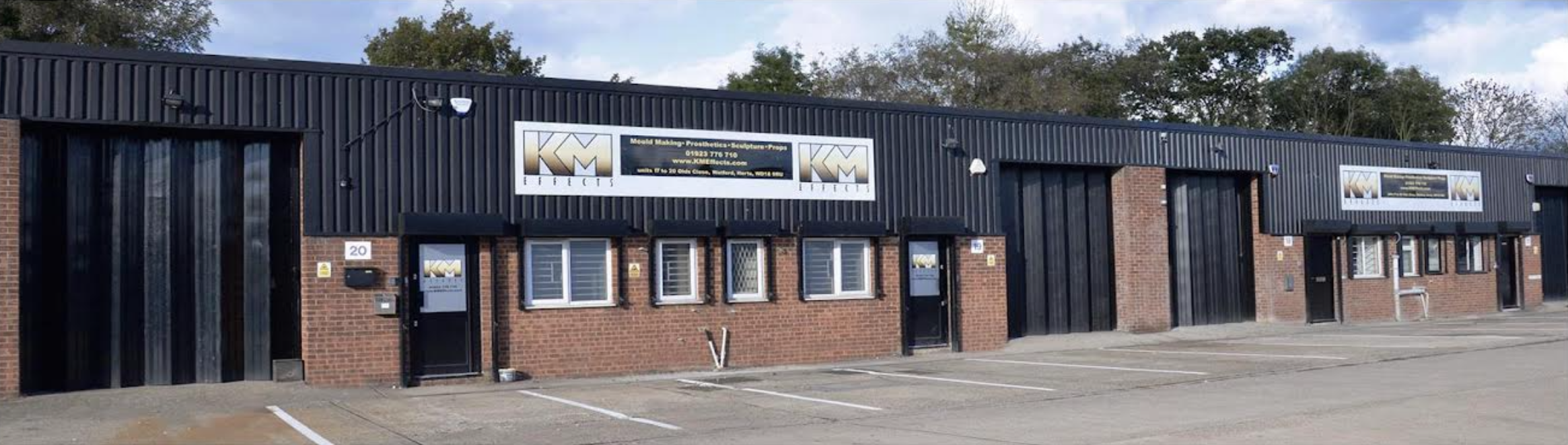 A row of industrial warehouse units is located here, with brick lower walls and black paneling above, featuring large garage doors, small windows, and KM Steelworks signs; the parking lot in front is mostly empty.